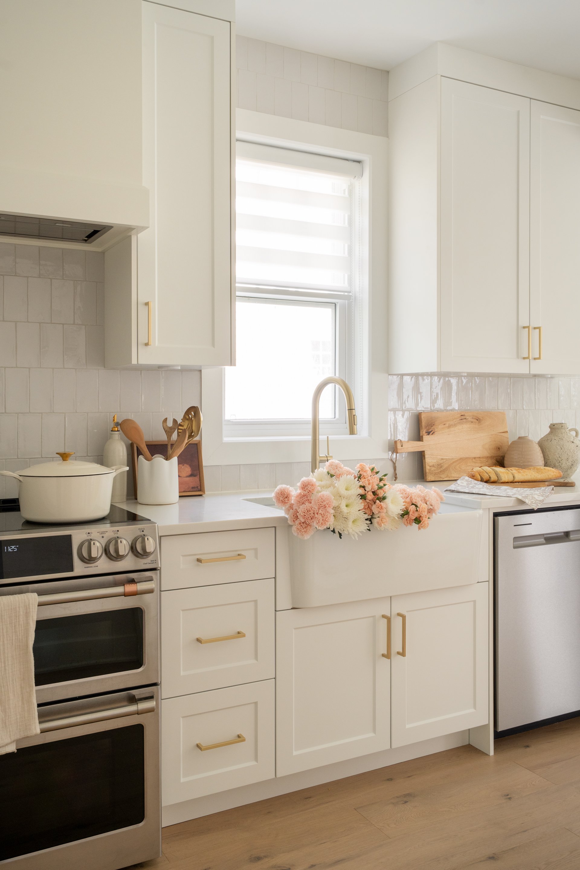 A bright white kitchen featuring a window with a flower arrangement in a farmhouse sink, white cabinets with gold handles, a stainless steel dishwasher, a stove with pots, and decorative items on the countertop.