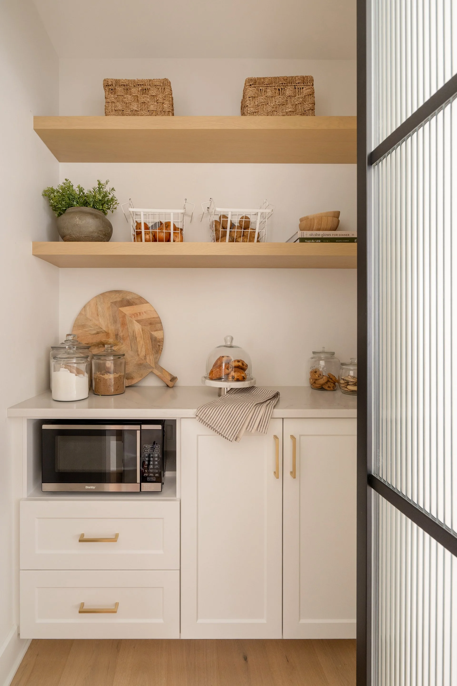 A small, white kitchen area with two shelves holding woven baskets, books, and small jars. The counter has jars of sugar, flour, and cookies, with a small microwave oven and a striped towel. The floor is wood, and part of a ribbed, black-framed glass wall is visible on the right.