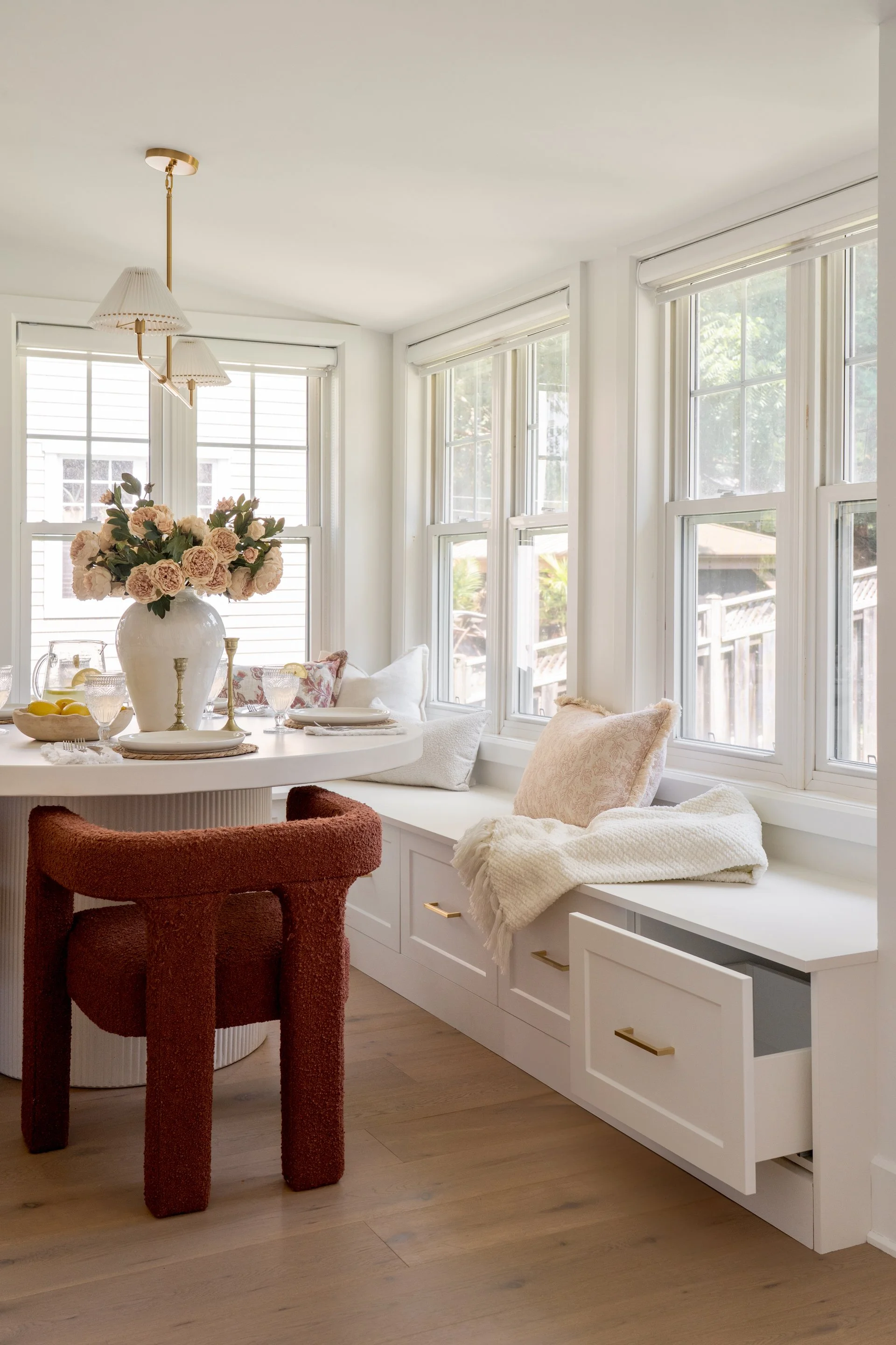 Sunlit breakfast nook with white built-in window bench, pillows, baskets, and a round table decorated with a large floral vase, plates, glasses, and lemons.
