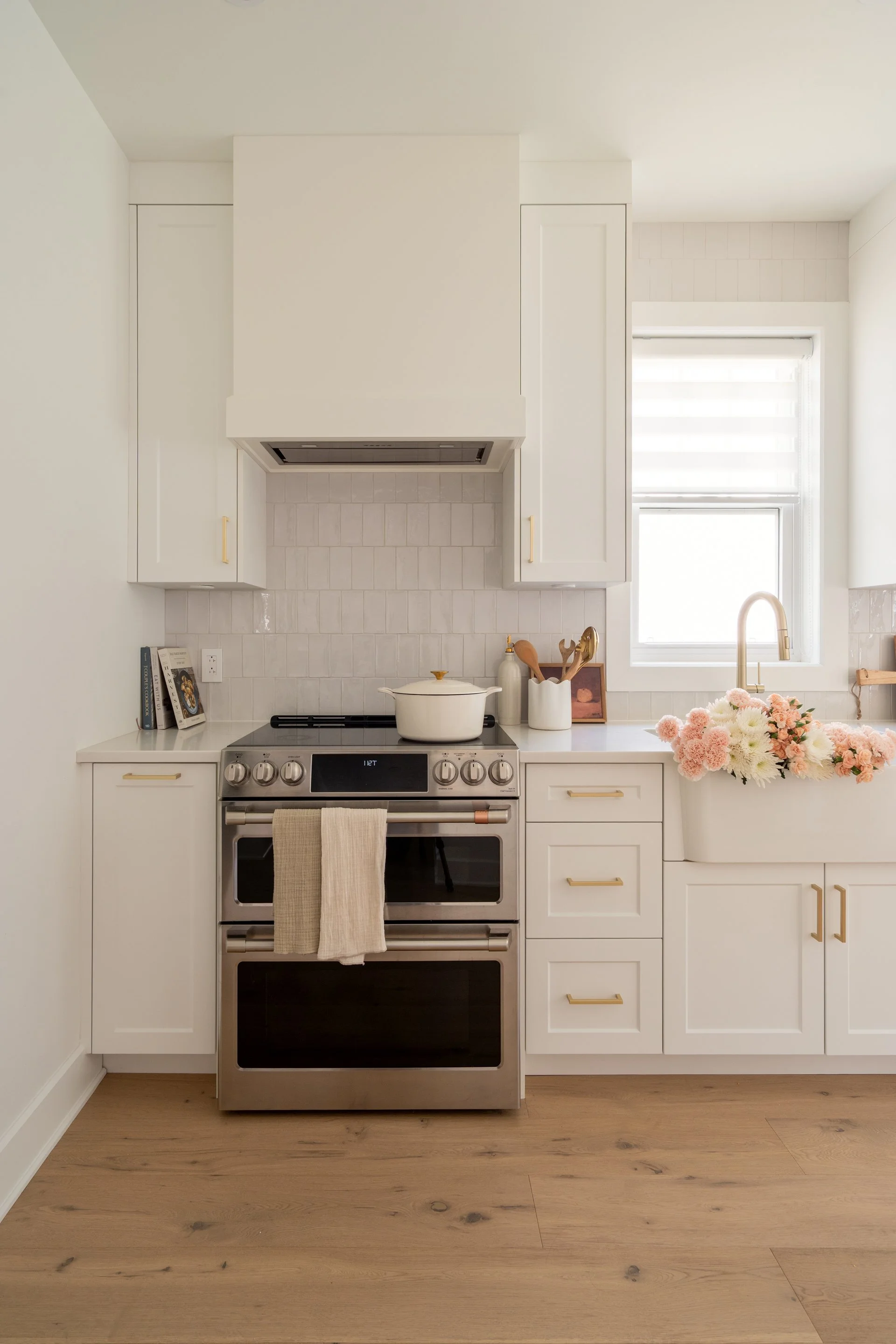 Modern white kitchen with stainless steel oven, a white pot on stove, a window with a white shade, and pink and white flowers on the sink