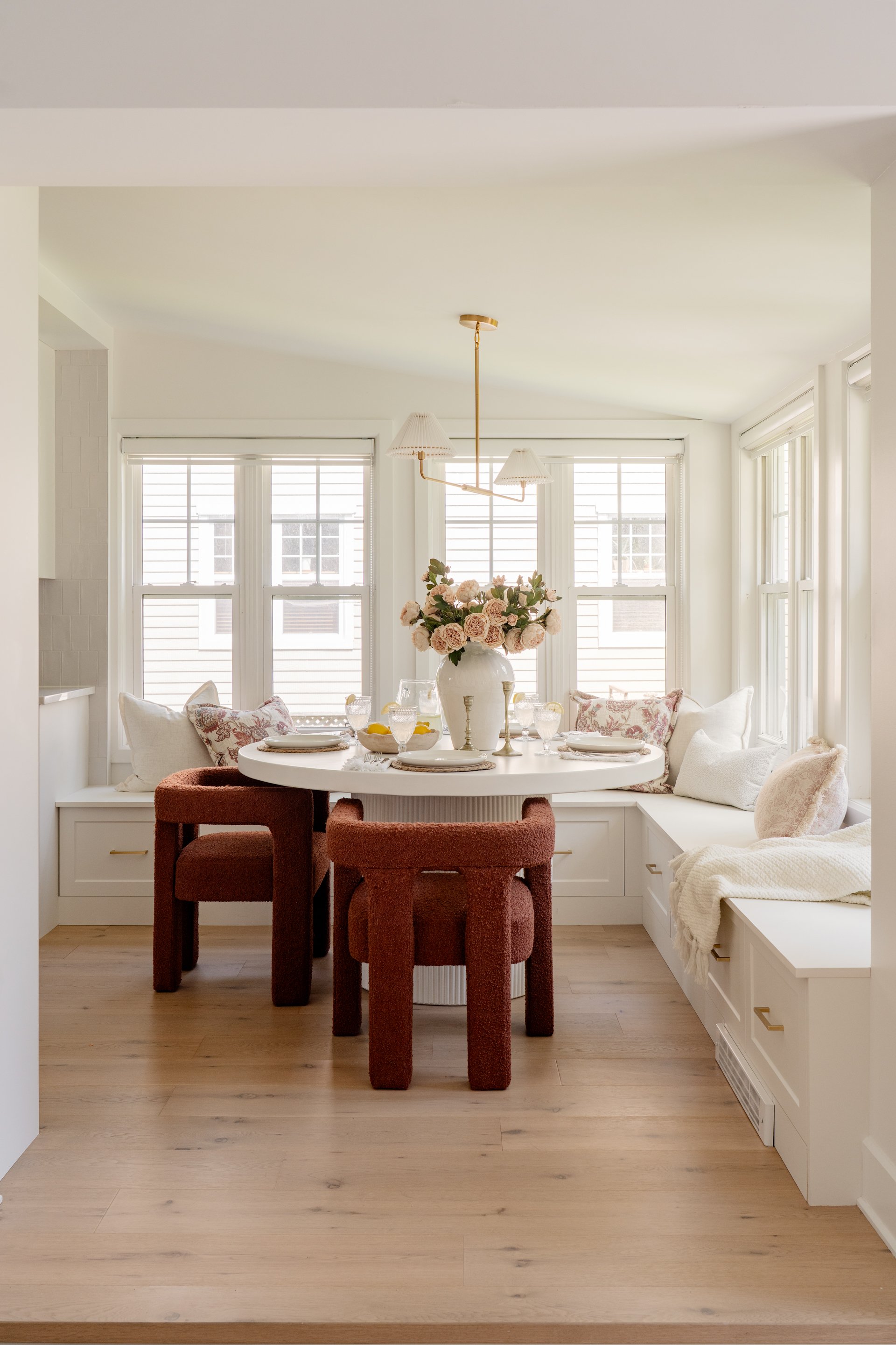 Bright dining area with round white table. Centerpiece with pink roses in a white vase. Four upholstered chairs, two rust-colored and two white, around the table. Built-in window seat with pillows. Multiple windows with white frames and shades. Light wood flooring.