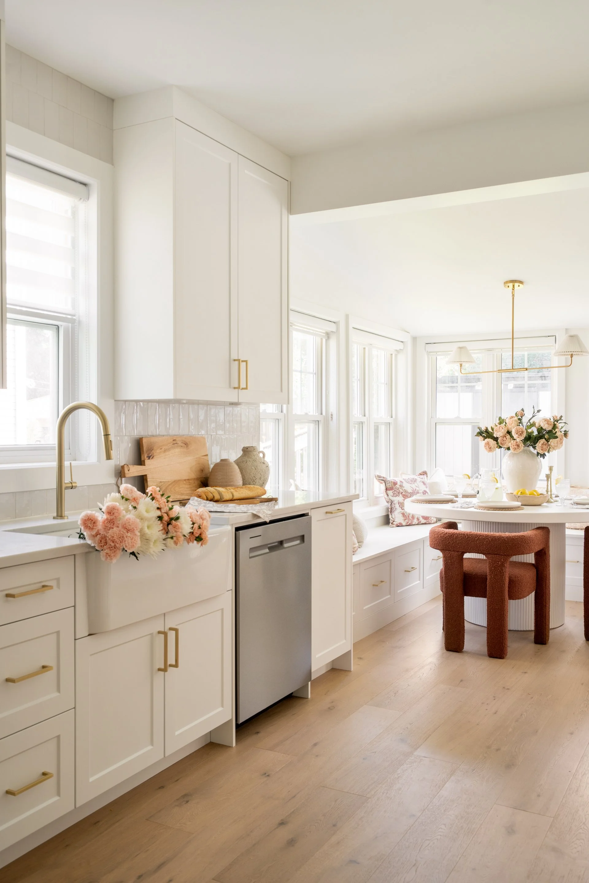 Bright kitchen with white cabinets, a window seat with pink floral pillows, a round table with a large pink flower arrangement, and a cozy brown chair. Light wood flooring and large windows let in natural light.
