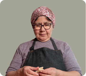 An older woman wearing glasses, a pink patterned headscarf, and a gray shirt, holding a small object in her hands, with a neutral background.