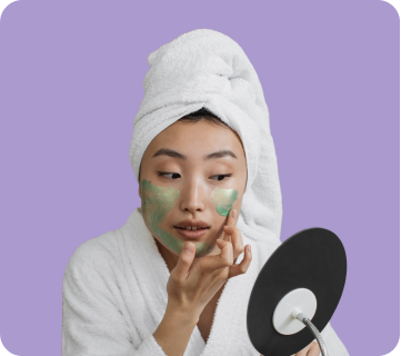 A woman with a towel on her head and facial masks applying cream to her face while looking in a mirror, against a light purple background.