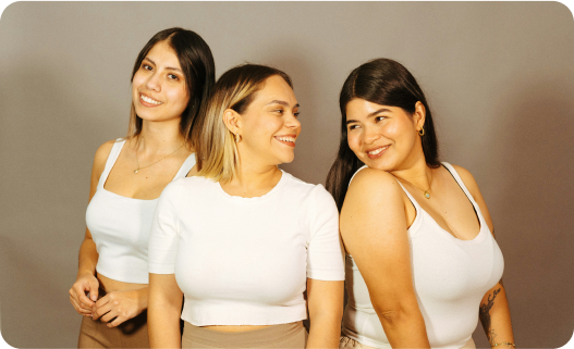 Three women smiling and looking at each other, standing against a neutral background, wearing white tops.