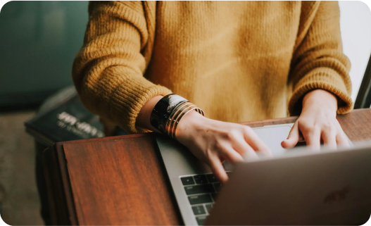 Person typing on a laptop at a wooden table with a yellow sweater and bracelets.