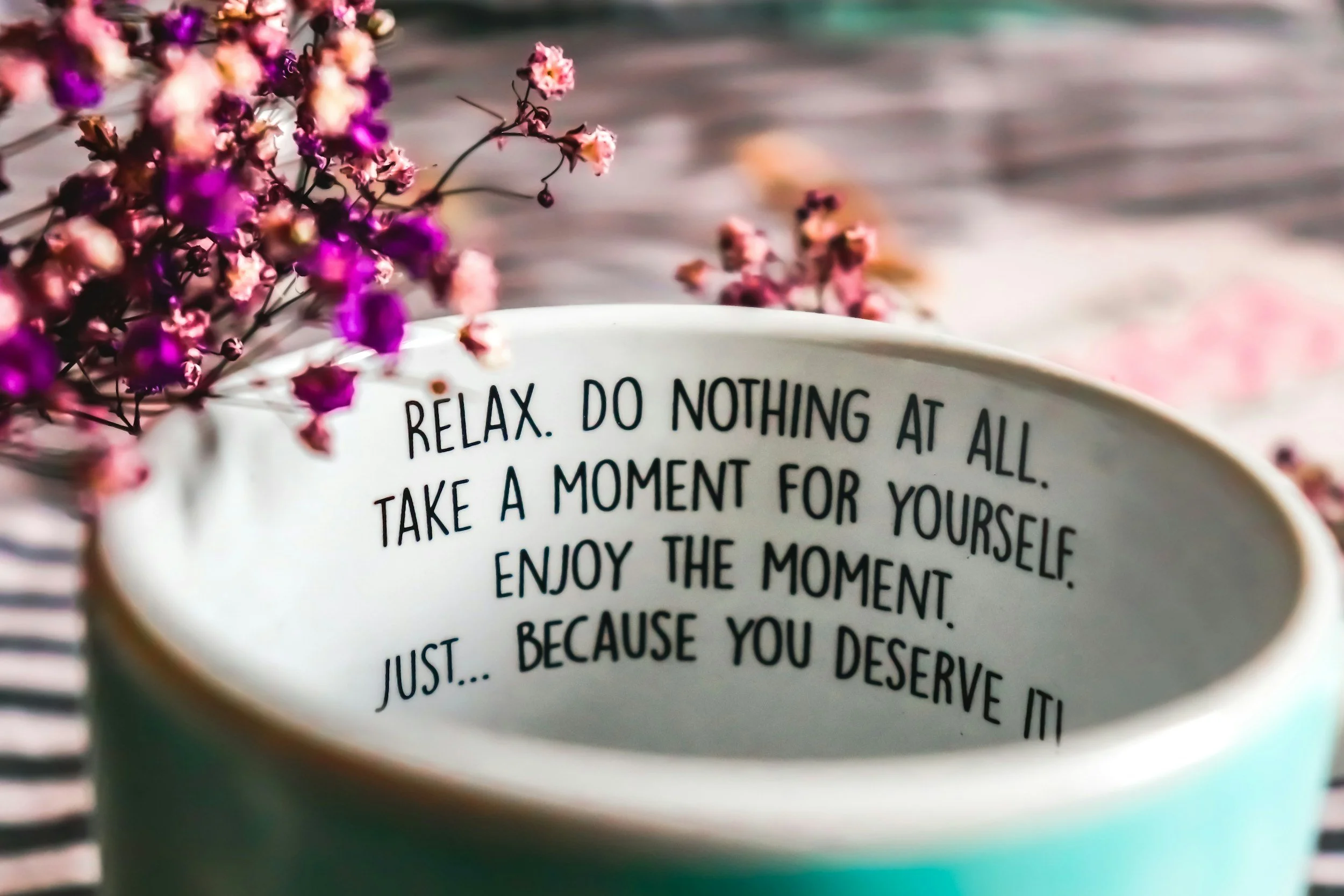 A white mug with a motivational quote inside, surrounded by pink and purple flowers, on a wooden surface.