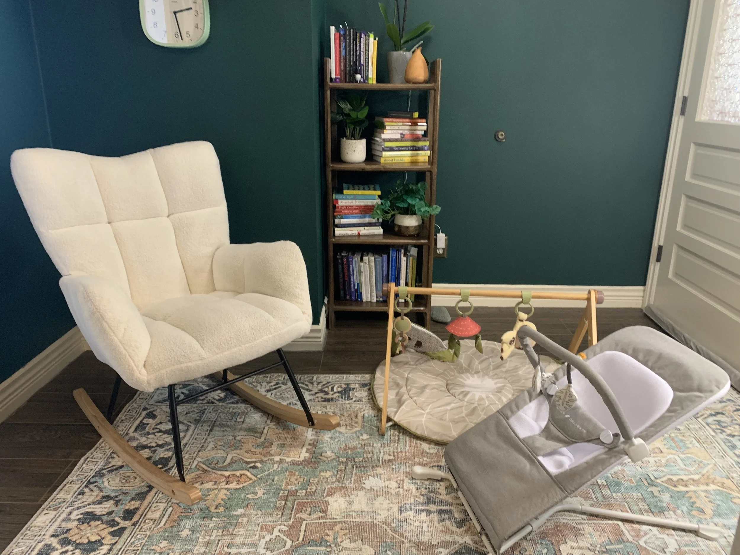 A cozy corner with a white rocking chair, a baby bassinet, a wooden bookcase filled with books, and a round activity mat with hanging toys, set on an oriental rug in a room with teal walls and a clock on the wall.