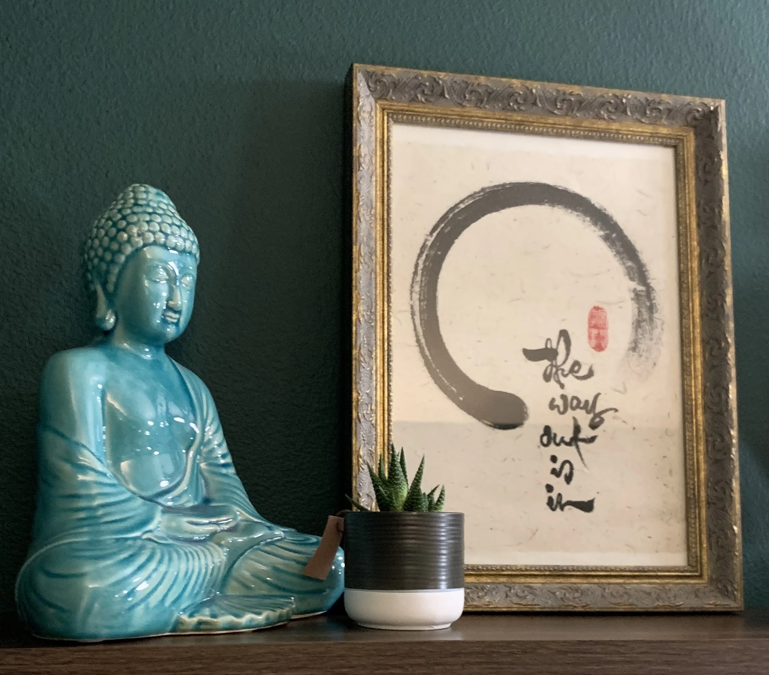 A turquoise Buddha statue with intricate details, a small potted succulent plant in a black and white pot, and a framed art piece with a black circle and handwritten text that reads "the war out is" on a dark green wall.
