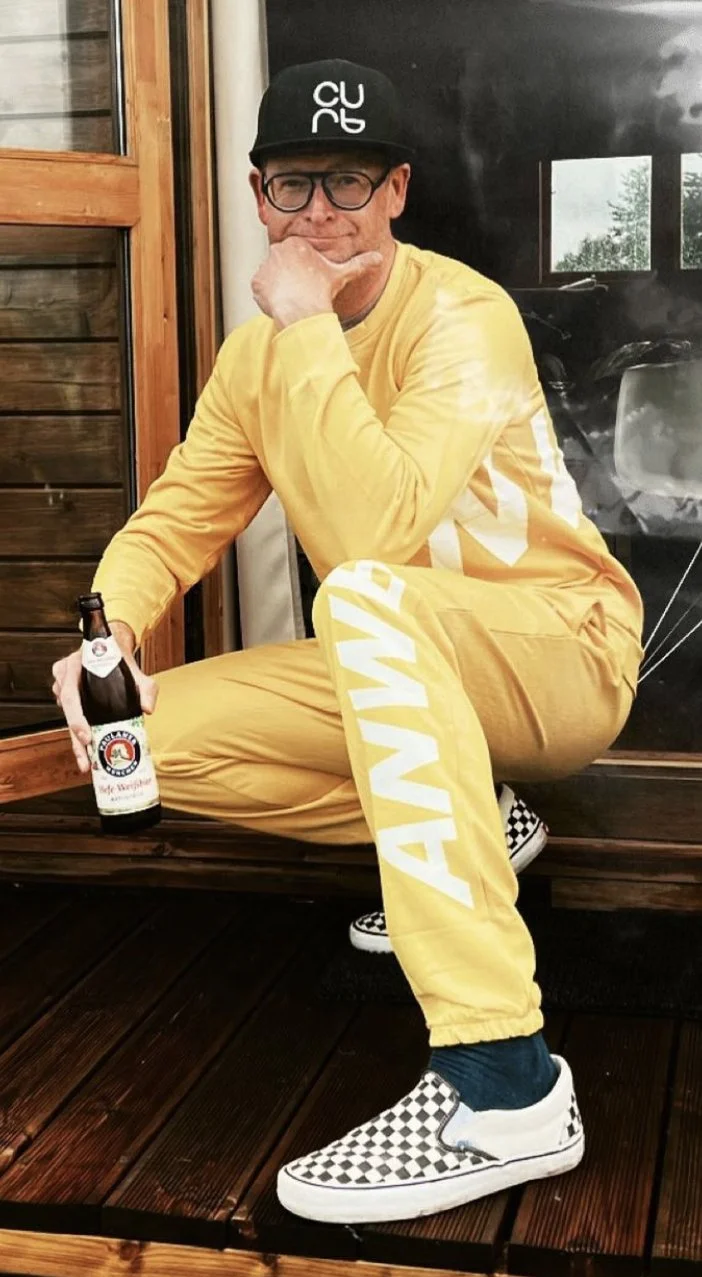 Mat in a yellow sweatsuit and black ball cap holding a beer while squatting down and smiling bravely at the camera
