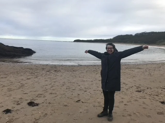Lisa standing on the beach in a black coat with arms outstretched looking confident