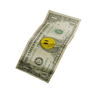 A one-dollar bill with a yellow smiley face sticker on it, placed on a light-colored surface.