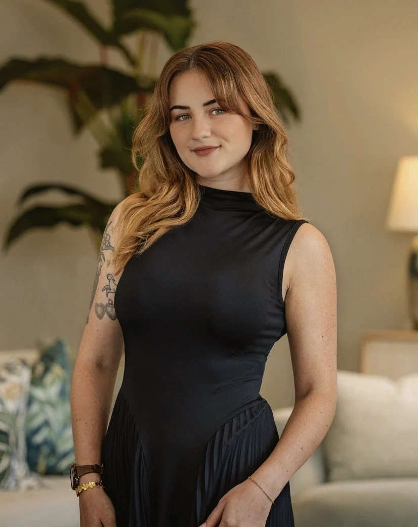 A young woman with wavy, shoulder-length brown hair and light skin standing in a warmly lit room. She is wearing a sleeveless black dress and has a tattoo on her upper left arm. She is smiling slightly and looking at the camera, with a background featuring a large plant, a lamp, and a bed.