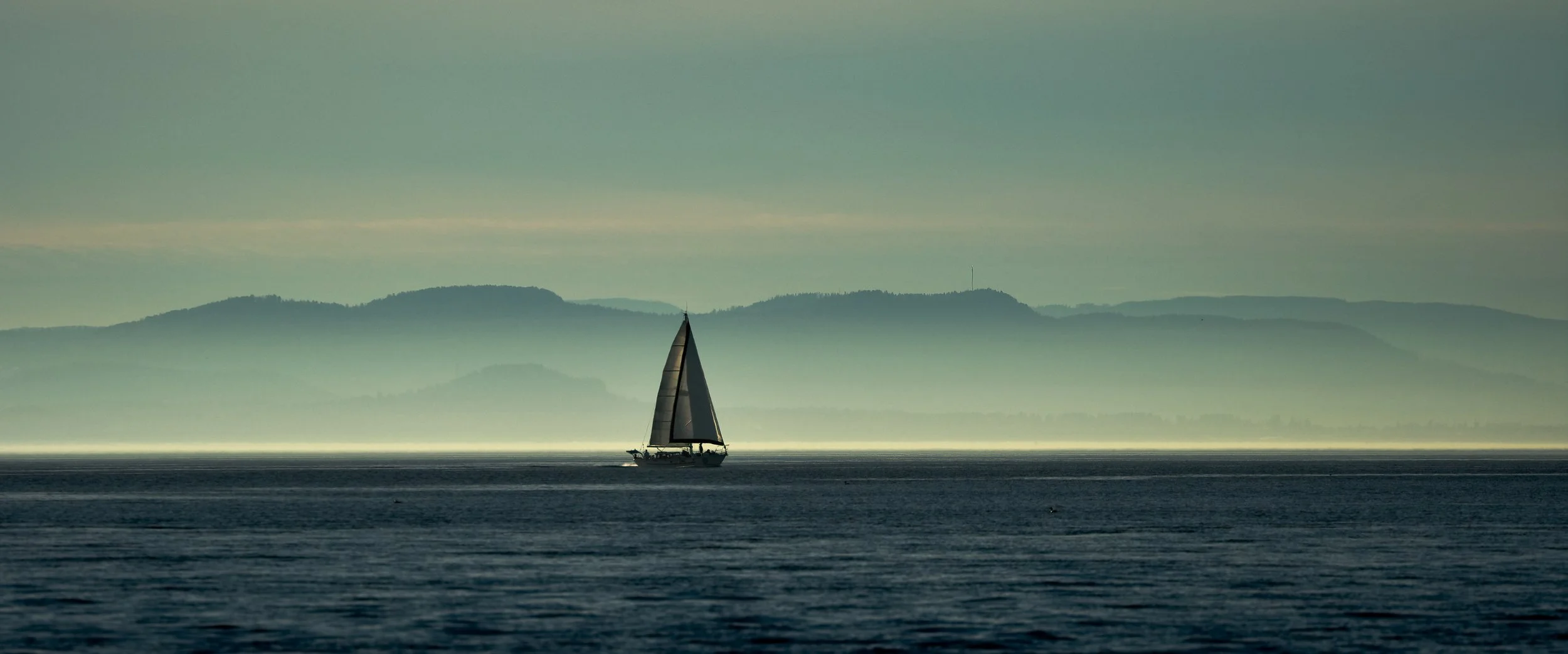 SAILING THE HARO STRAIT – The misty mountains of Sidney Island, Canada create a perfect backdrop for a passing sailboat.