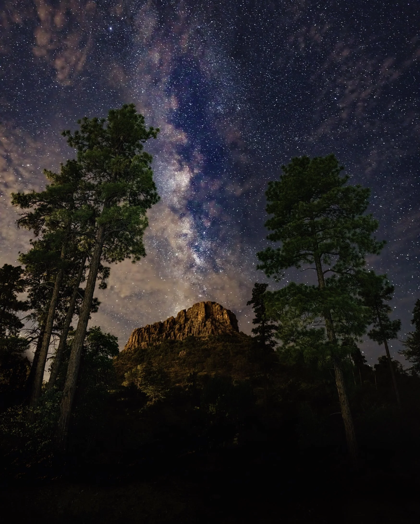 CELESTIAL PINES – Thumb Butte is the signature geological feature in Prescott, Arizona. I wanted to capture The Milky Way towering vertically above the mountain as if it were a volcanic eruption of stars. Thumb Butte was illuminated by the city light