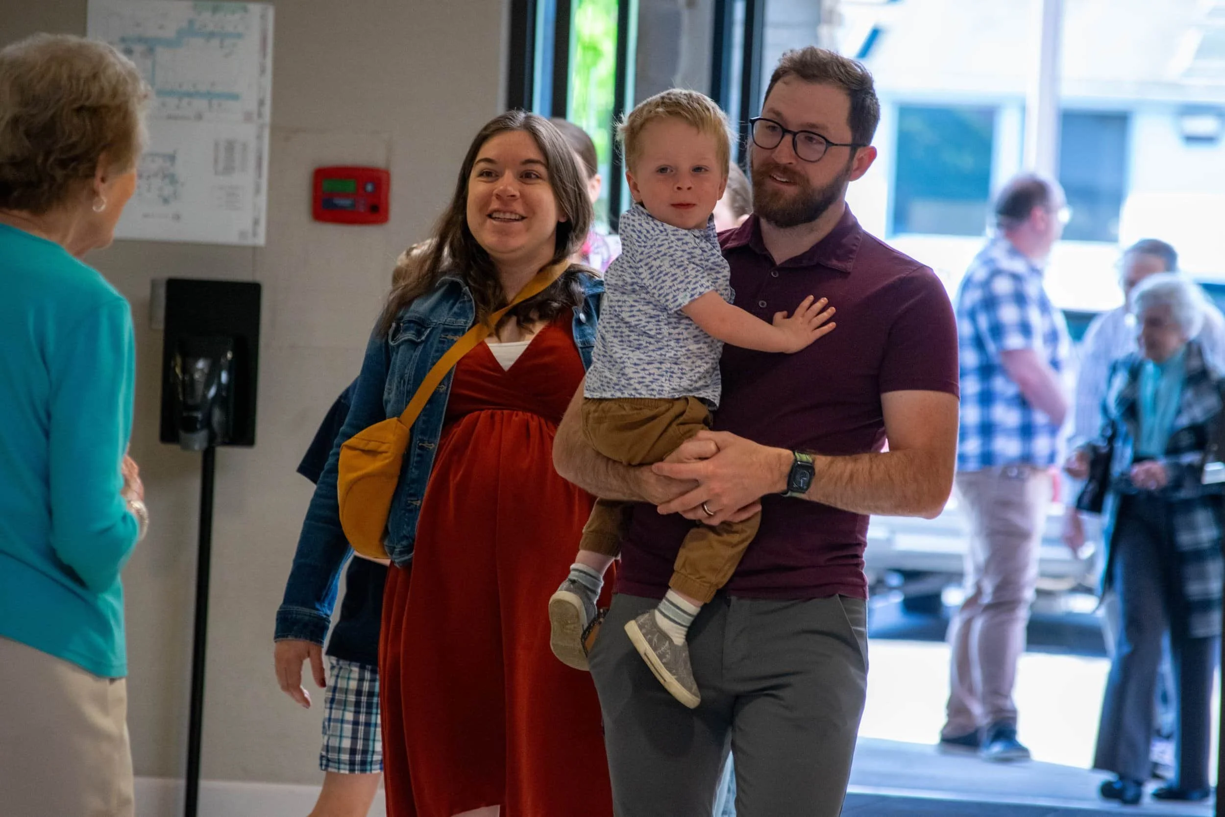Man holding young boy with pregant mom next to them walking through the lobby of church with an elderly person greeting them and more people being greeted and entering the building through the doors in the background.