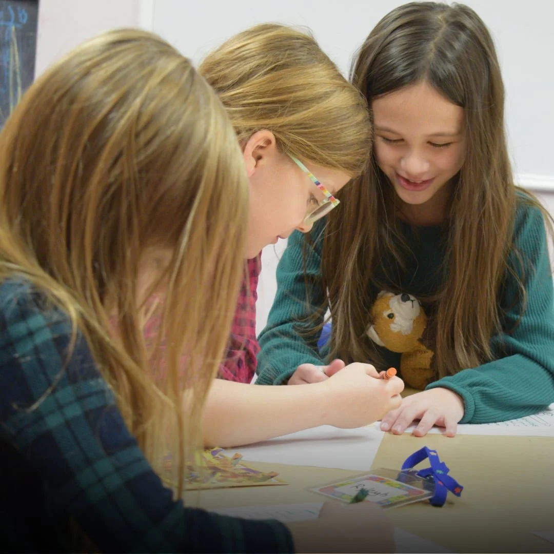 Three young girls with long hair, two with light brown and one with dark brown, gathered around a table, engaging in a storytelling activity, smiling and focusing on the papers in front of them.