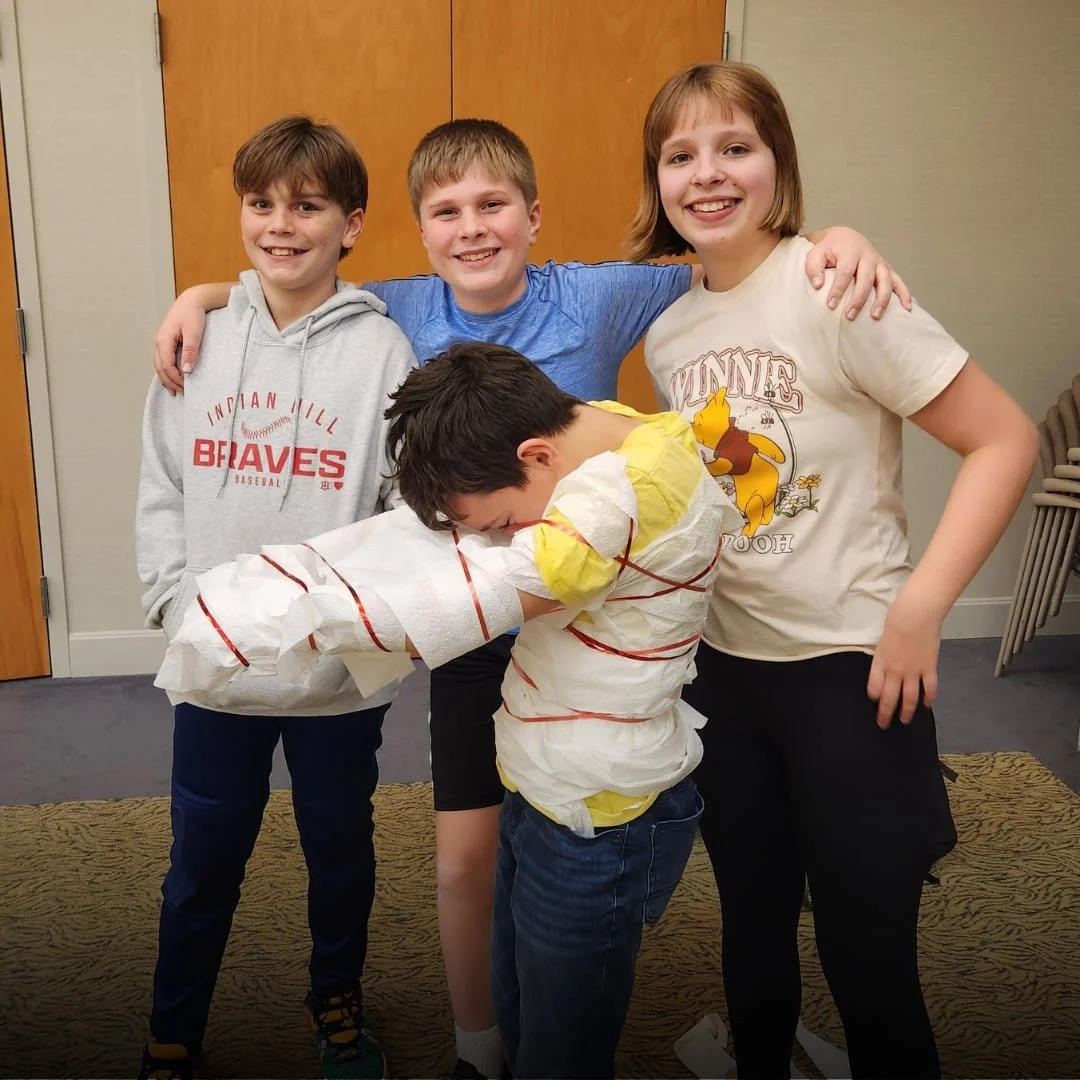 Four children standing together, with one boy wrapped in toilet paper, pretending to be a mummy, while the other three kids smile and pose with him.