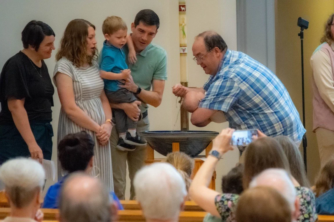 A pastor baptizing a baby during worship at a baptismal font with parents and a sibling excitedly overlooking the ceremony. Baptismal candle in the background of image.