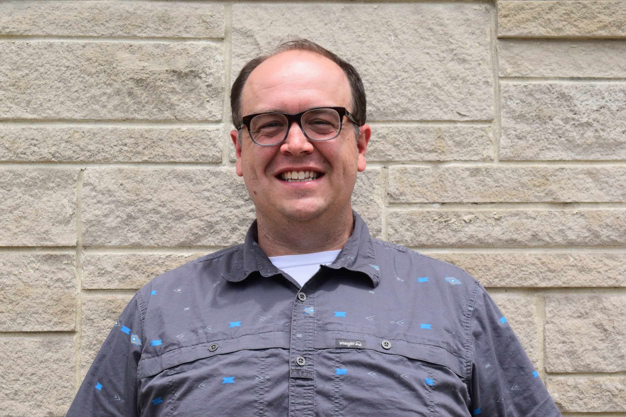 A smiling man with glasses standing in front of a beige brick wall.