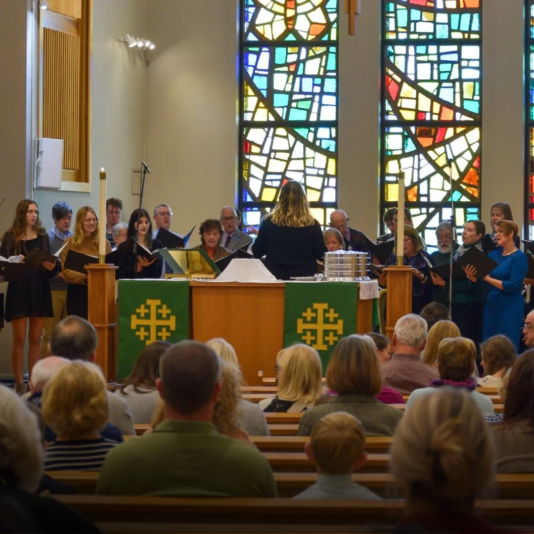 People singing in the church choir with leader conducting them in the foreground and a stained glass window in the background.