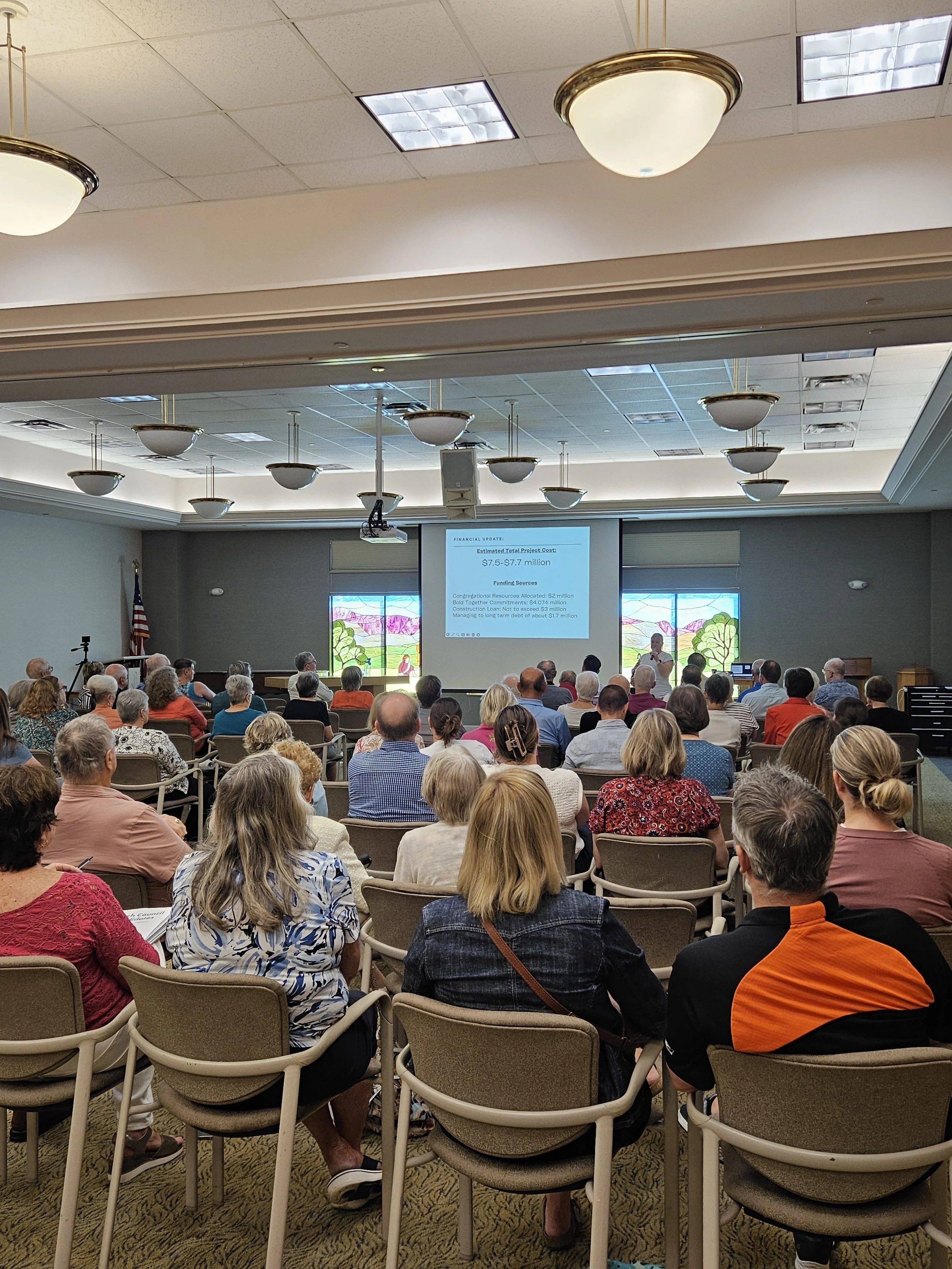 A large gathering of people attending a presentation in a conference room. The presenter is at the front, standing near a screen displaying a financial update and project costs. The audience is seated facing the speaker, with stained glass windows behind. The ceiling has multiple hanging light fixtures.