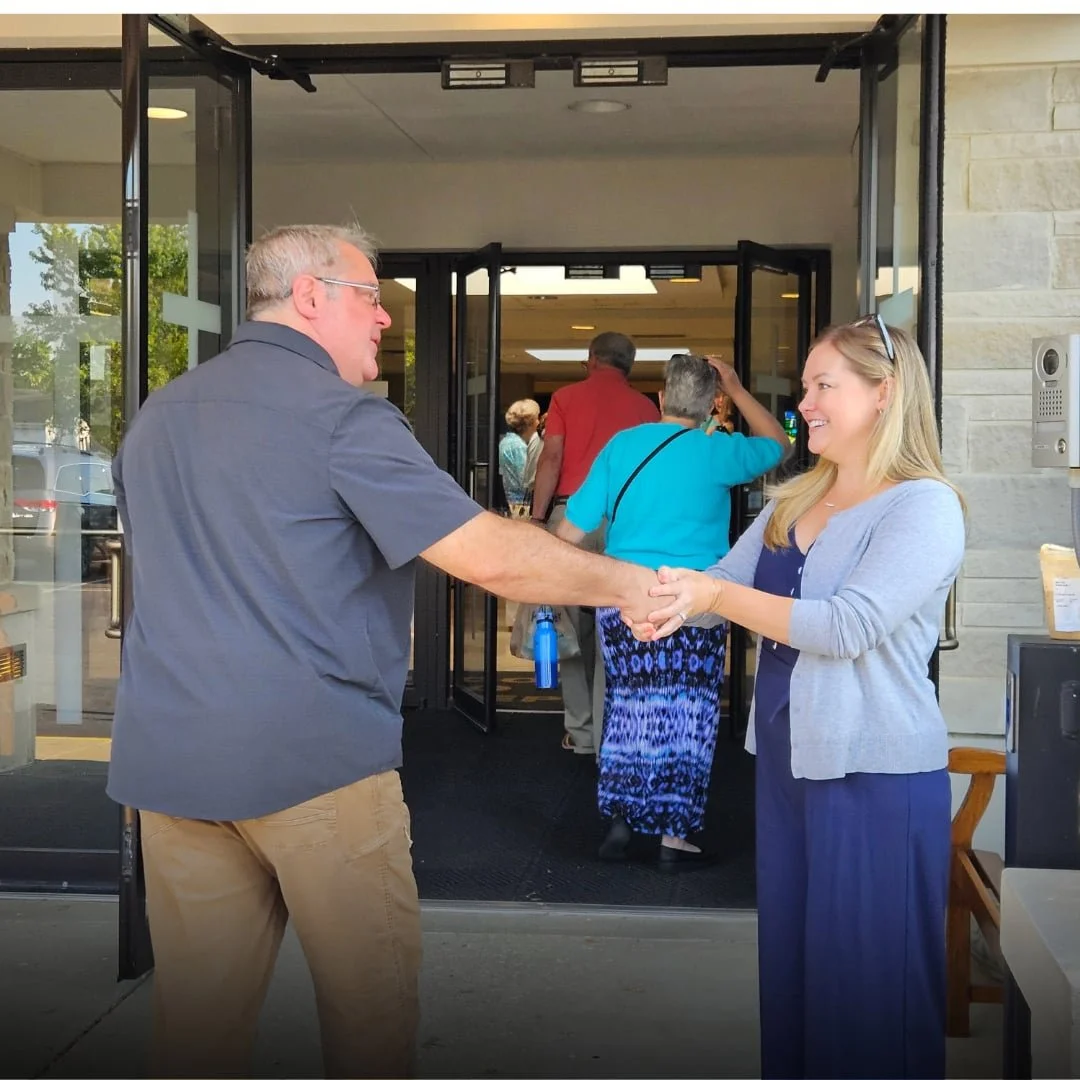 Welcome Team volunteer shaking hands with a person heading into the building with entrance doors in the background.