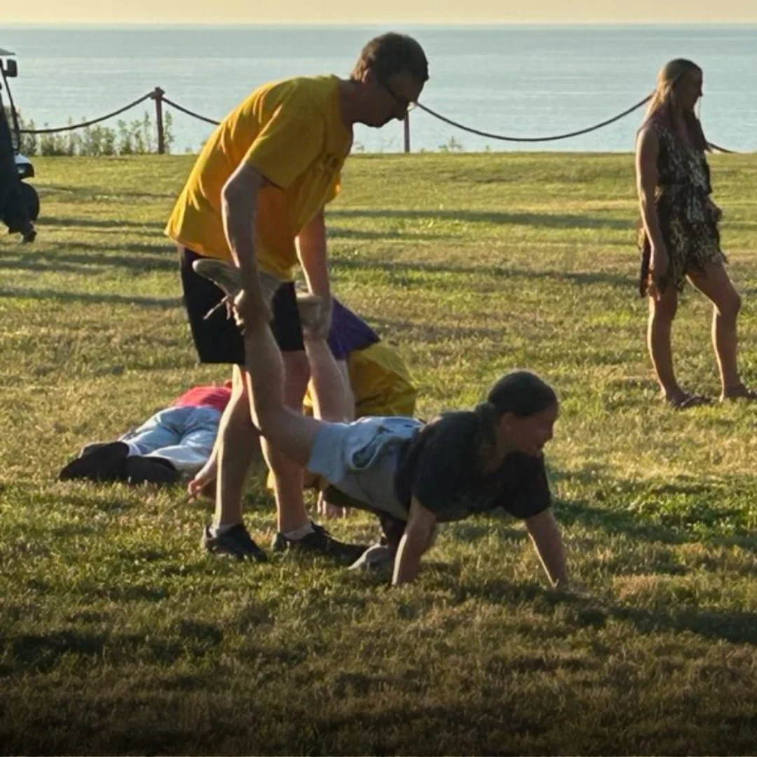Two people having fun at family camp outdoors on grass near water, with other people in the background.