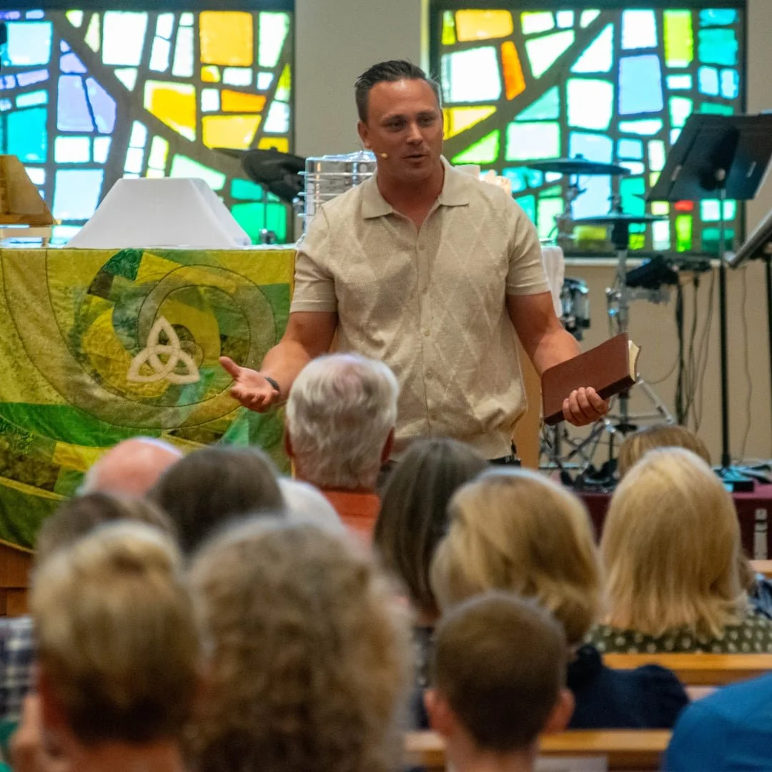 Picture of a pastor preaching during worship in front of an altar and stained glass window with worshipers in the foreground.
