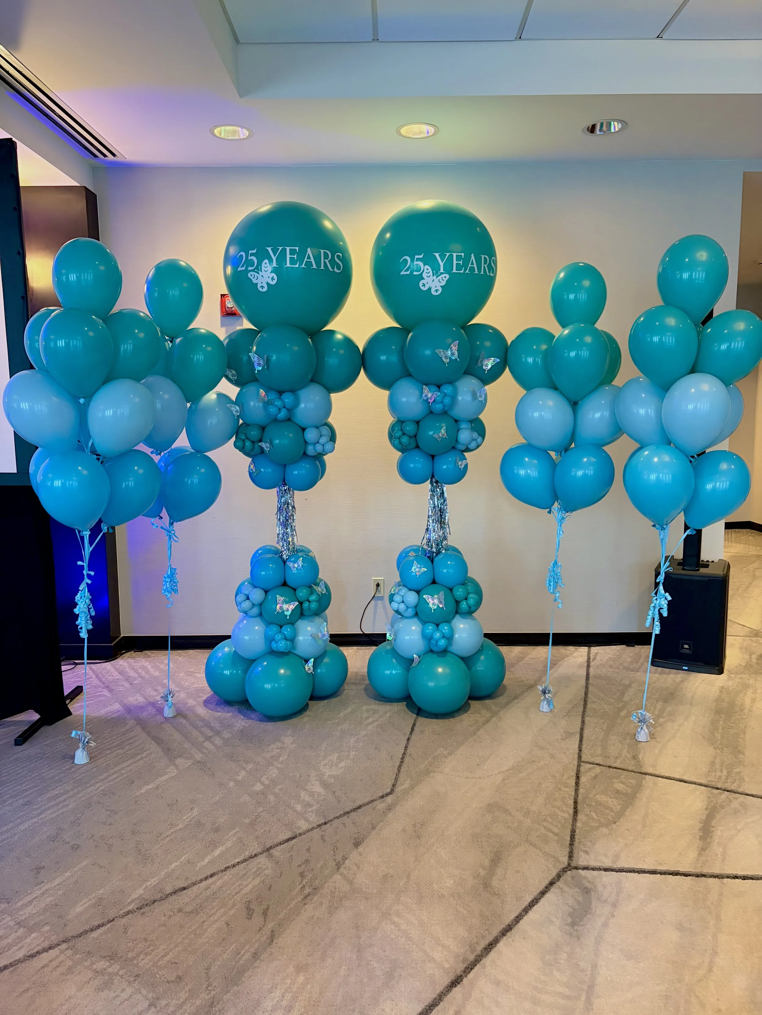 A decorative display of blue balloons celebrating a 25th anniversary or milestone. The arrangement features large balloons with '25 YEARS' and butterfly designs, supported by balloon sculptures with smaller balloons and butterfly embellishments, all set up indoors.