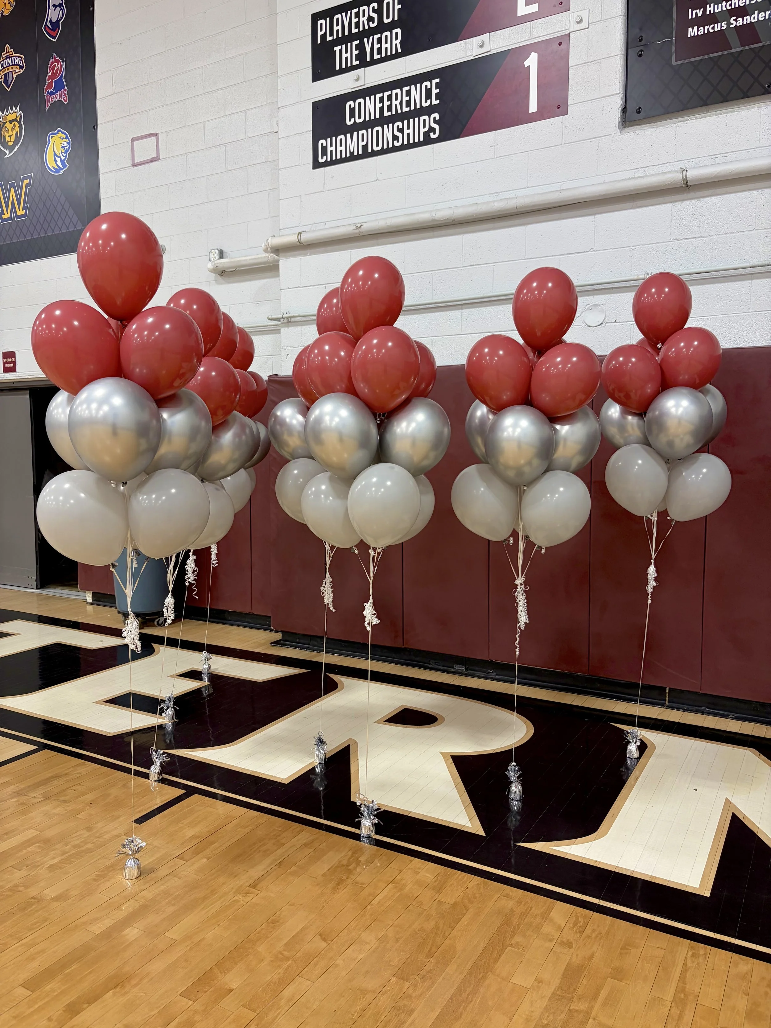 Group of red, silver, and white balloons arranged on a basketball court, with a banner displaying sports team logos and a sign indicating conference championships in the background.