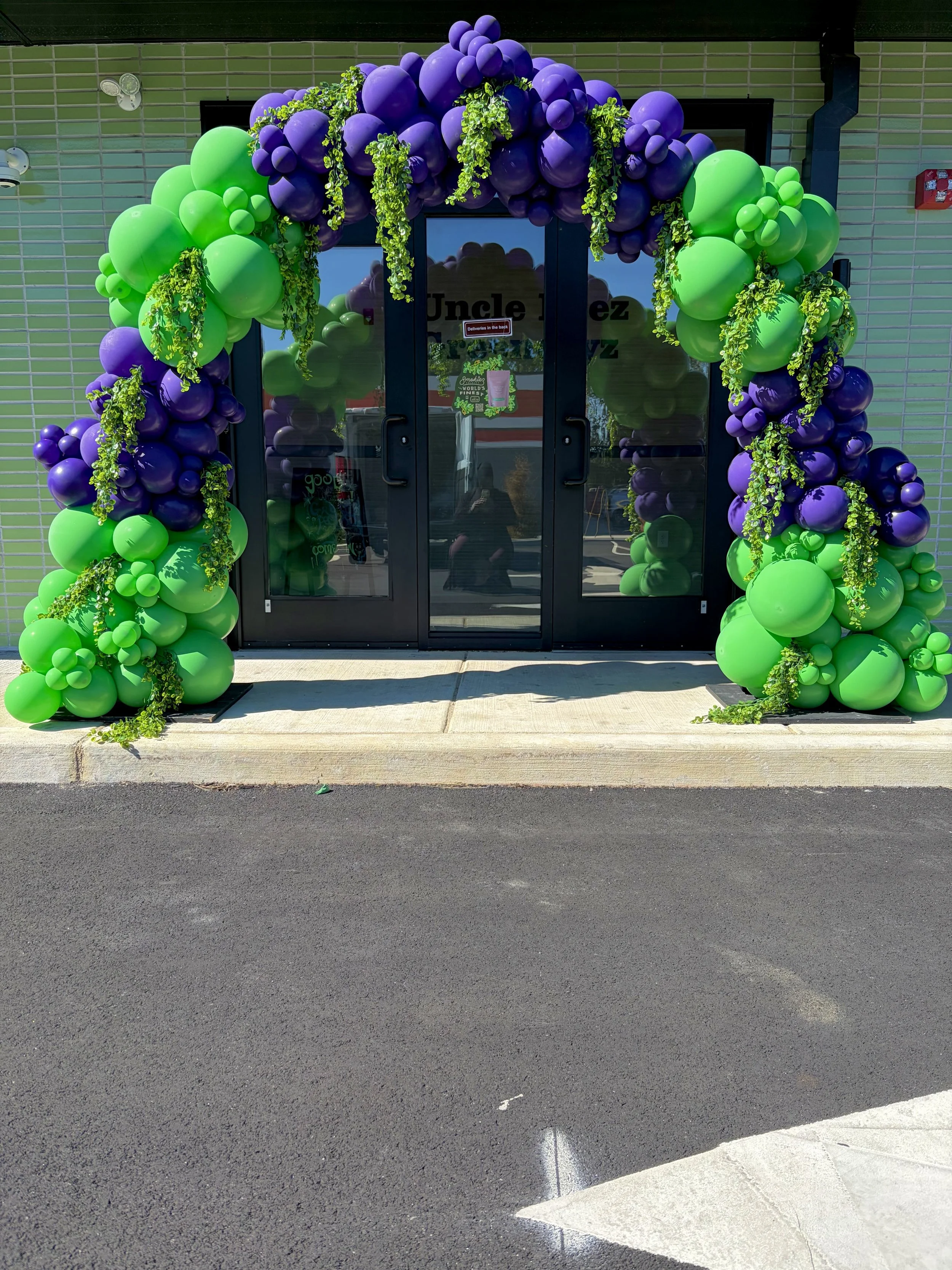 Colorful balloon arch with purple and green balloons and green greenery decoration at a building entrance.