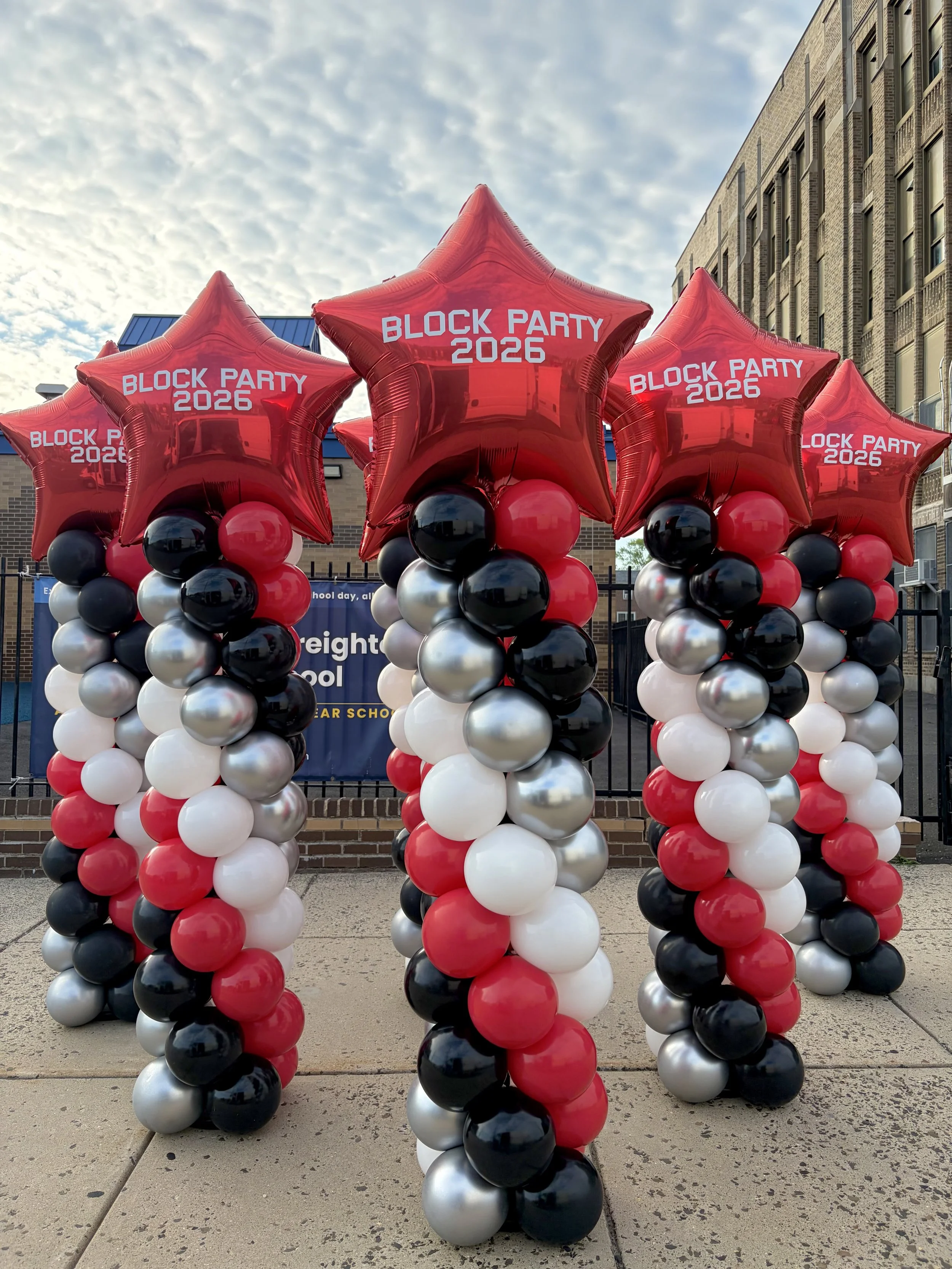 Red star-shaped balloons with white text reading 'BLOCK PARTY 2026', surrounded by black, white, and silver balloons, arranged in columns outdoors with a brick building and cloudy sky in the background.