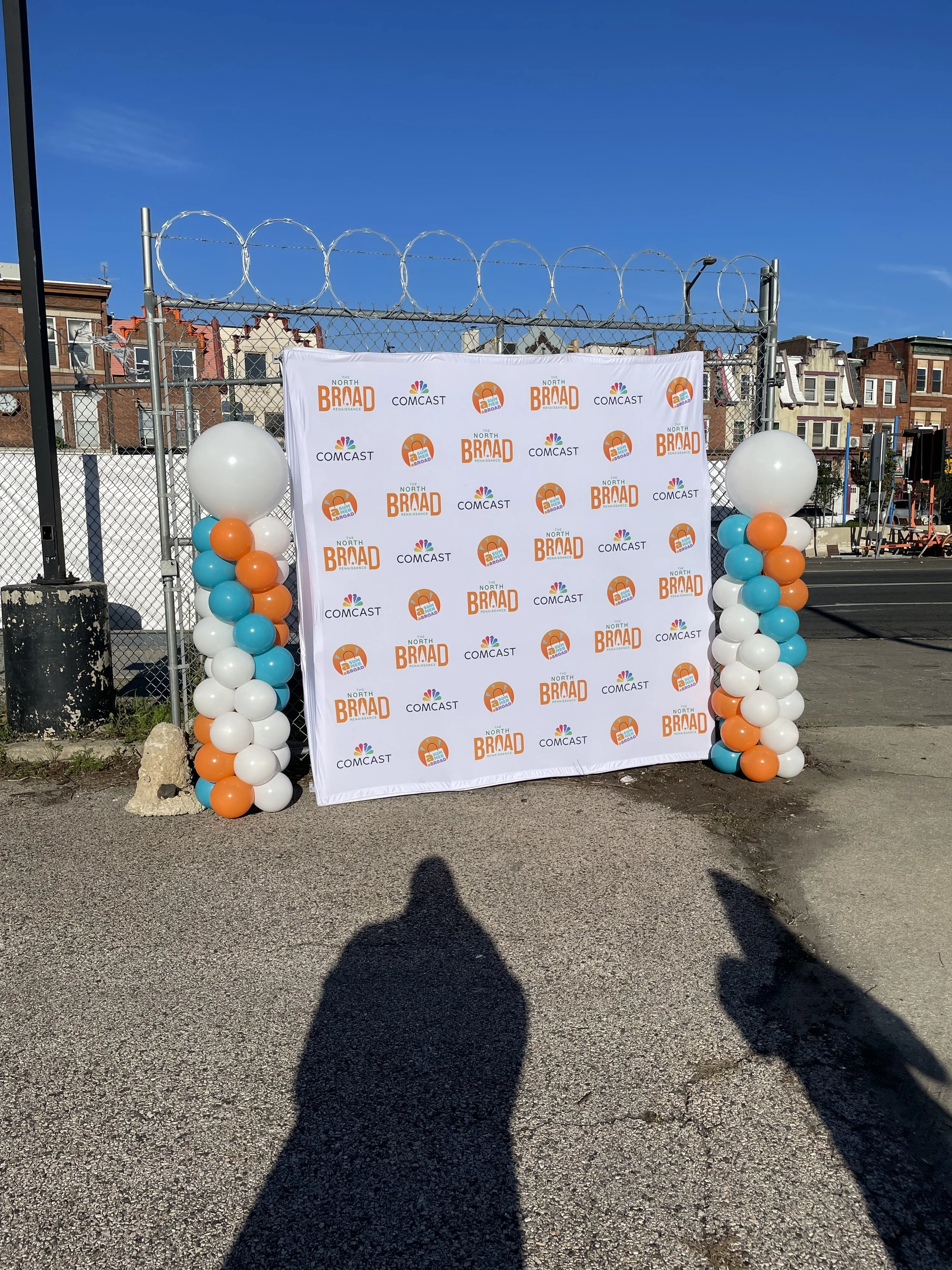 A step and repeat banner with logos of 'The North Broad Renaissance', 'Comcast', and an orange logo with the words 'A Sun for All'. The banner is decorated with columns of balloons in white, orange, and blue, and is placed in an outdoor setting with buildings in the background and a clear blue sky.