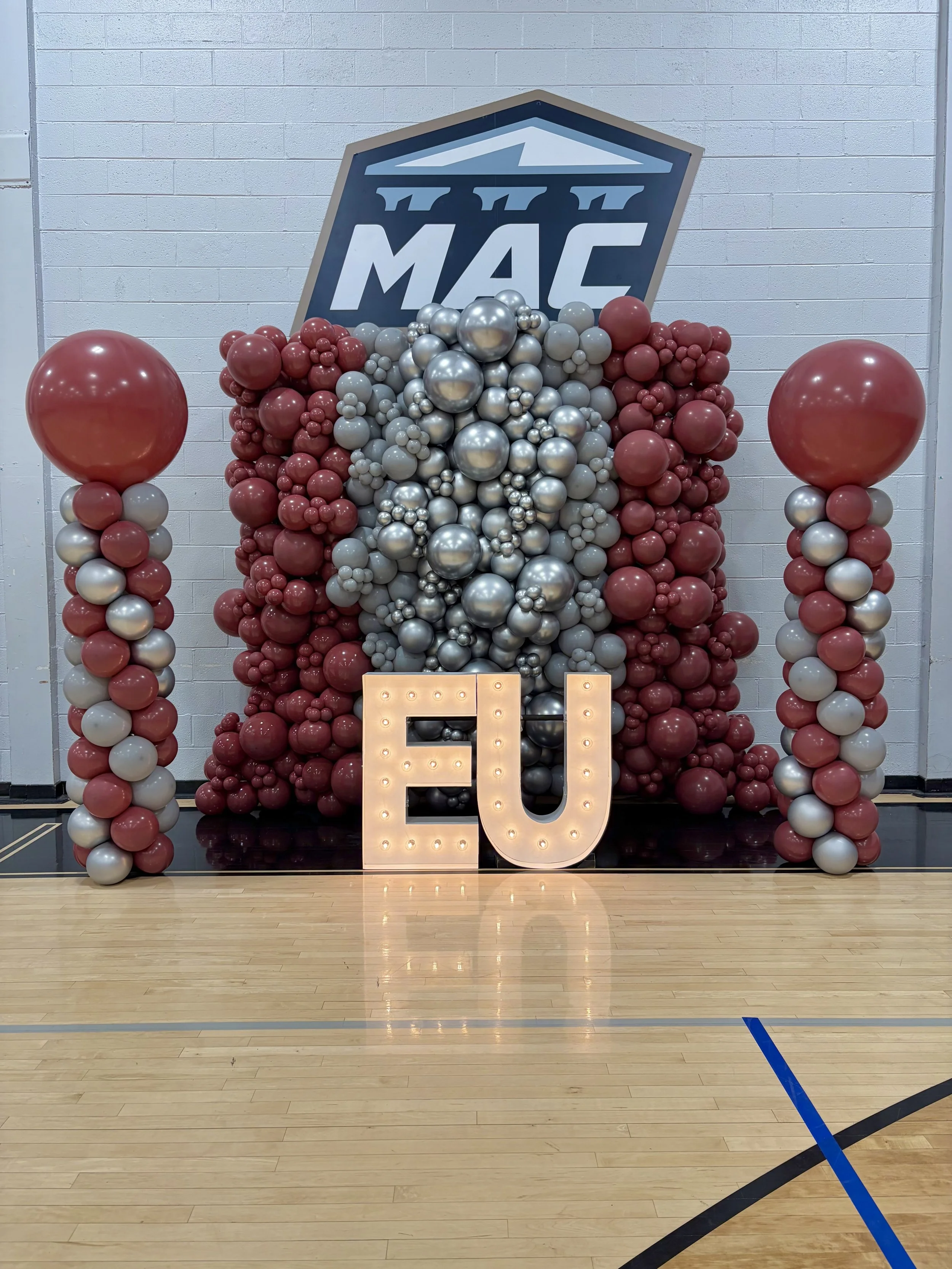 A basketball court decorated with a large arrangement of red, gray, and silver balloons, and illuminated marquee letters spelling 'EU.' A logo featuring a Greek temple with the word 'ΜΑΕ' in Greek letters is displayed in the background.