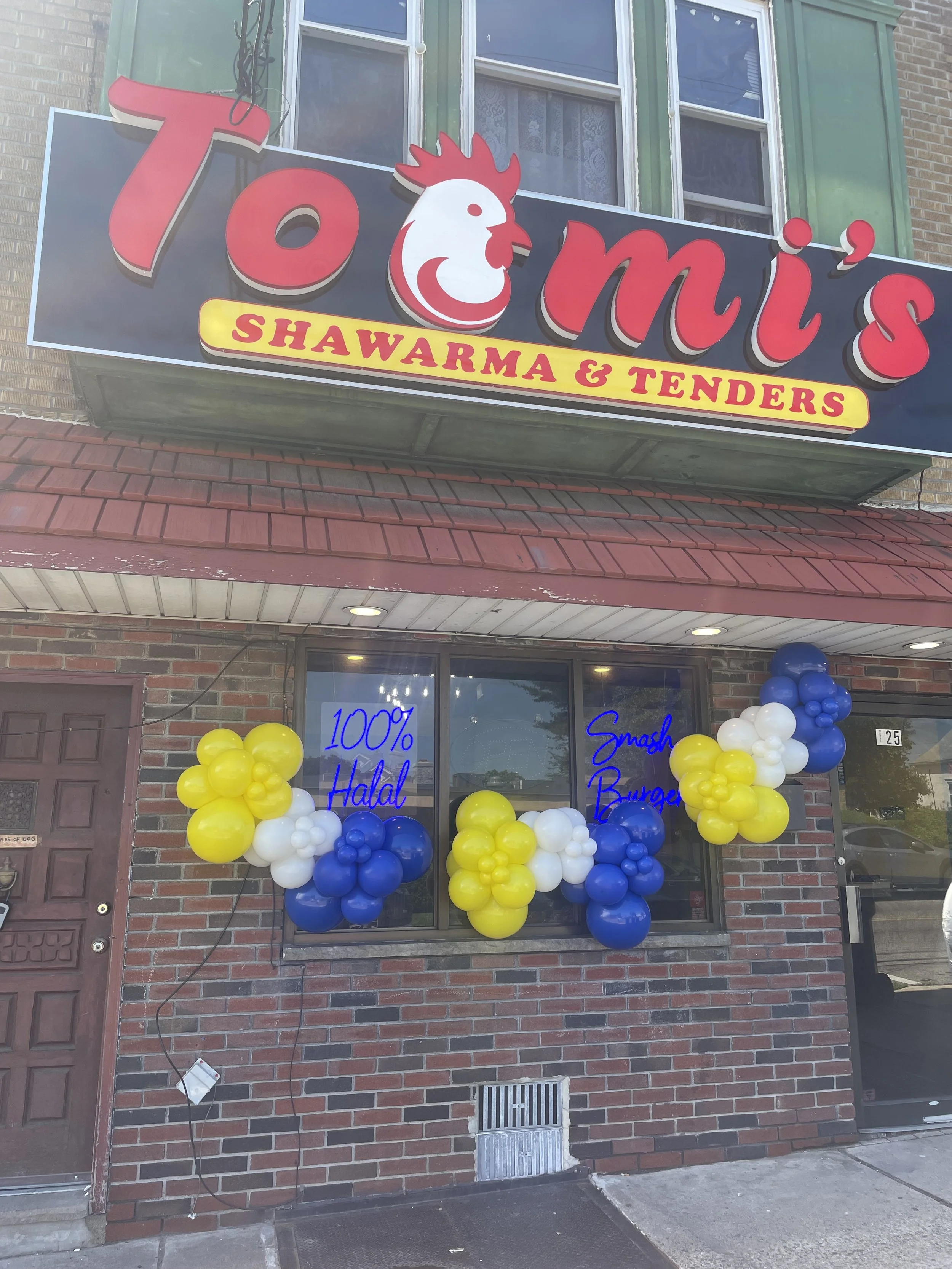 The storefront of a restaurant with a sign that reads 'Tomi's Shawarma & Tenders' with a cartoon chicken logo, decorated with blue, white, and yellow balloons, and windows with writing on them.
