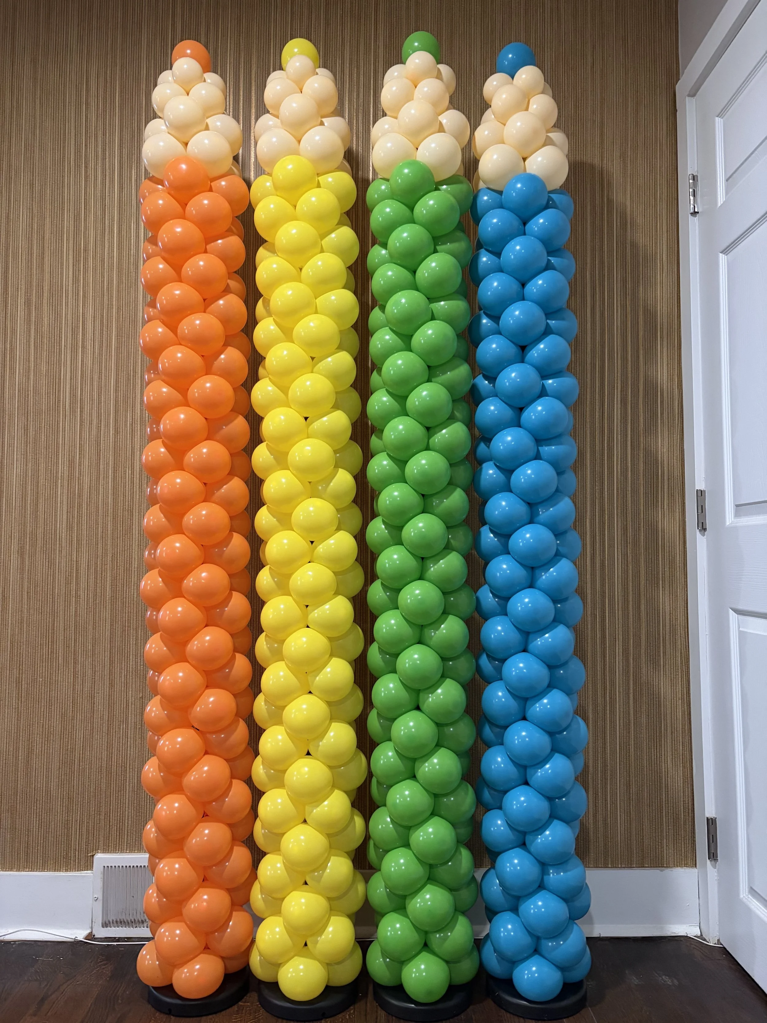 Four columns of balloons arranged in rainbow colors: orange, yellow, green, and blue, set against a brown wooden wall with a white door and baseboard.