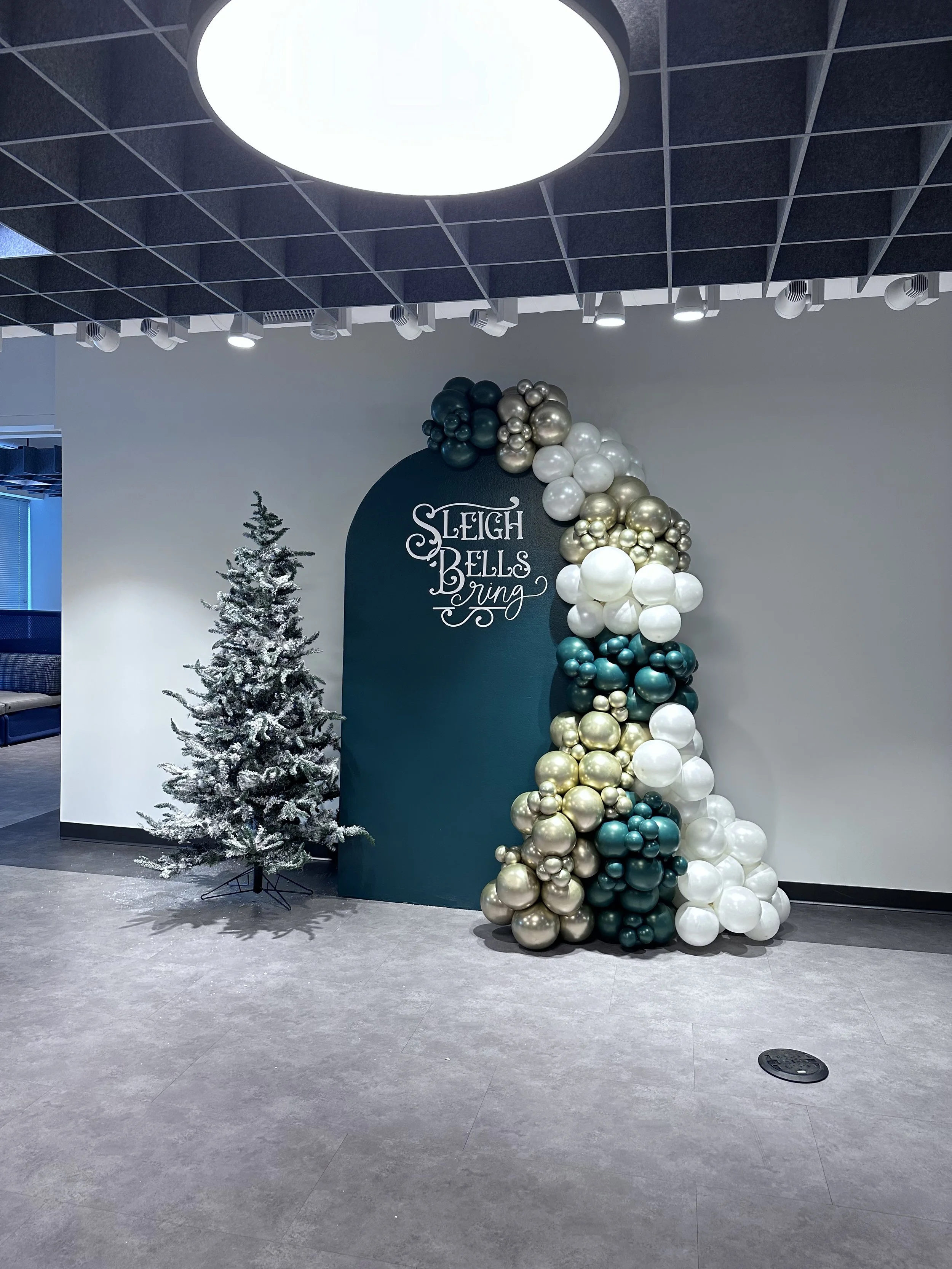 Decorative Christmas display with a snow-flocked Christmas tree and a large balloon arrangement in metallic colors, adjacent to a teal arch with festive lettering, in an indoor setting with a gray tile floor and a drop ceiling.