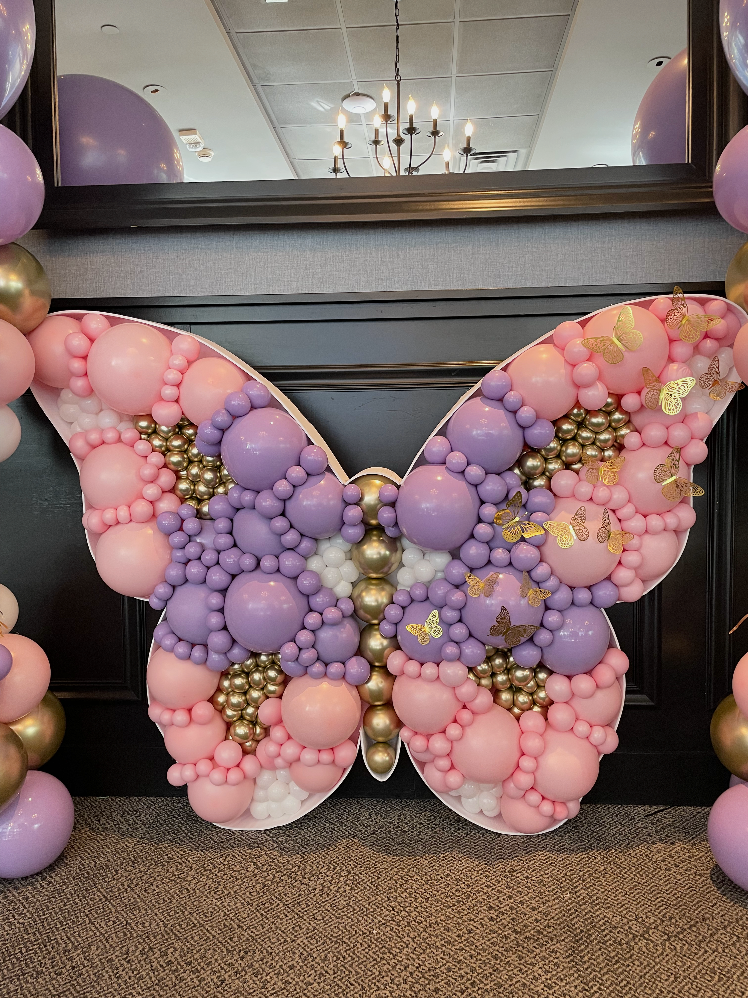 Large pink and purple balloon butterfly with gold and printed yellow butterflies on the wings, surrounded by smaller balloons, set against black wall panels and a carpeted floor, with a mirror and ceiling light fixture visible in the background.
