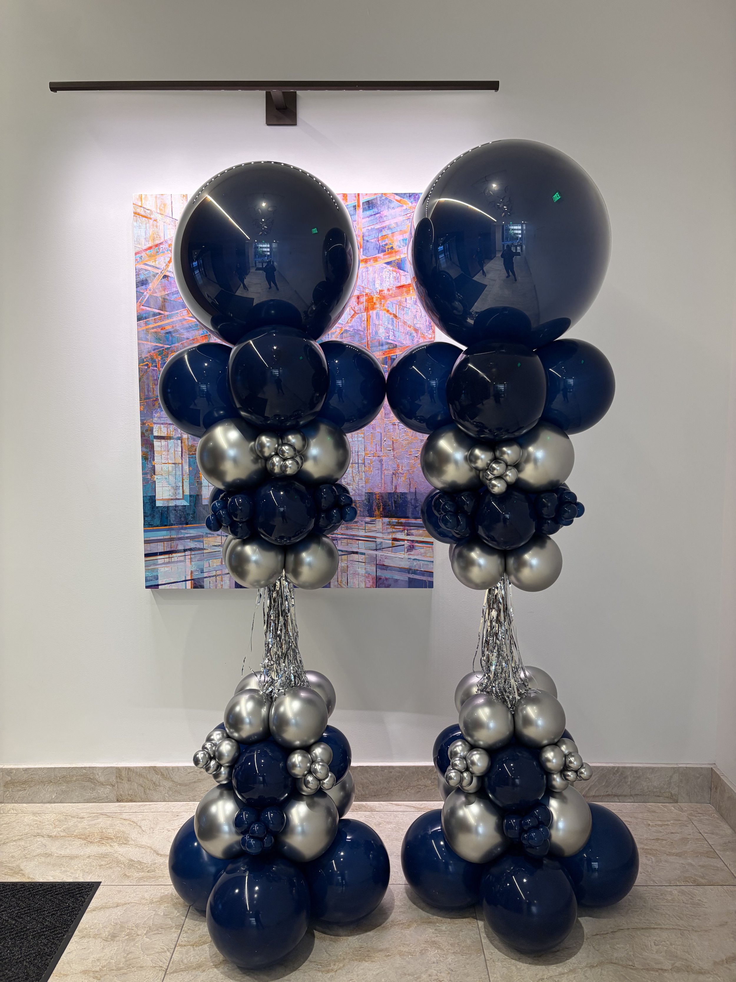 Two balloon columns made of navy blue, silver, and smaller black balloons, topped with large black reflective balloons, set against an abstract colorful painting, in an art gallery.
