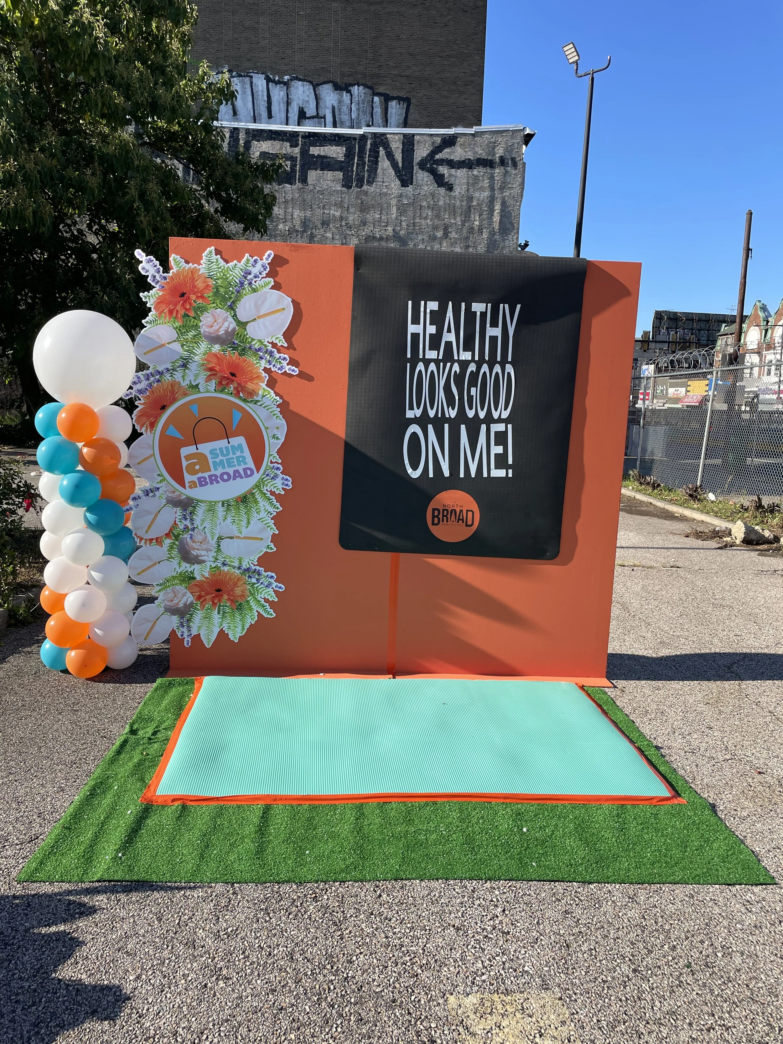 Outdoor photo of a sign that says 'Healthy Looks Good On Me!' with a floral decoration, balloons on the side, and a green and orange mat on the ground, under a clear blue sky.