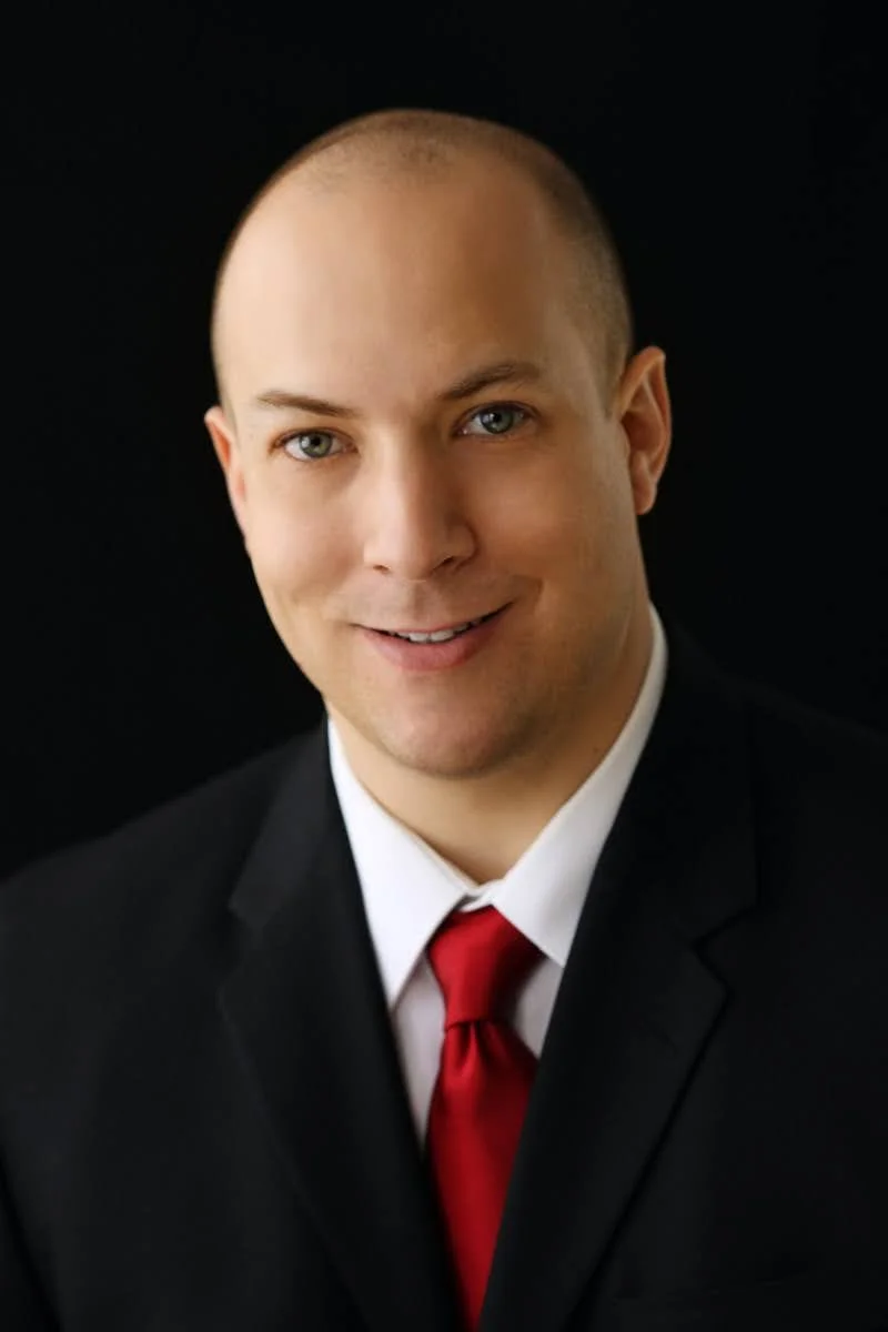 Portrait of a smiling man in a black suit, white shirt, and red tie against a black background.