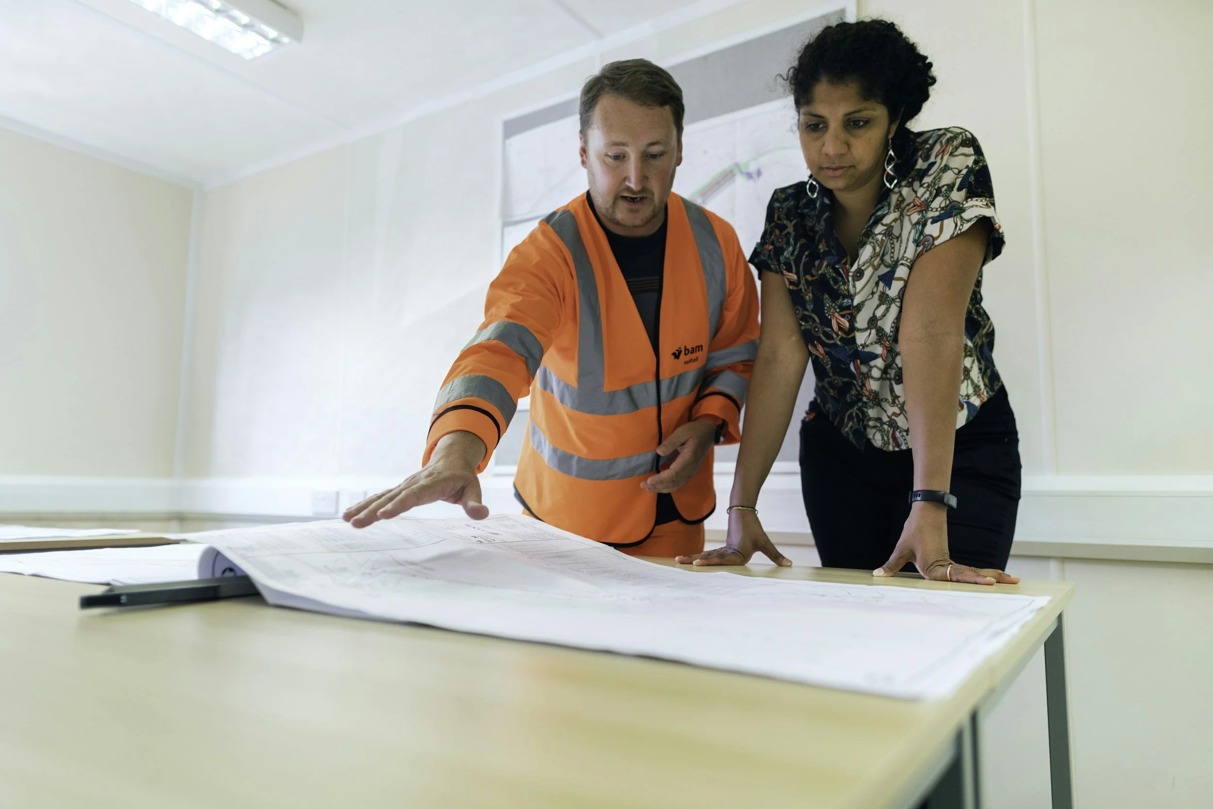 Two professionals, one male construction worker in an orange safety vest and one woman in casual clothing, reviewing large plans or blueprints on a table in an office or meeting room.
