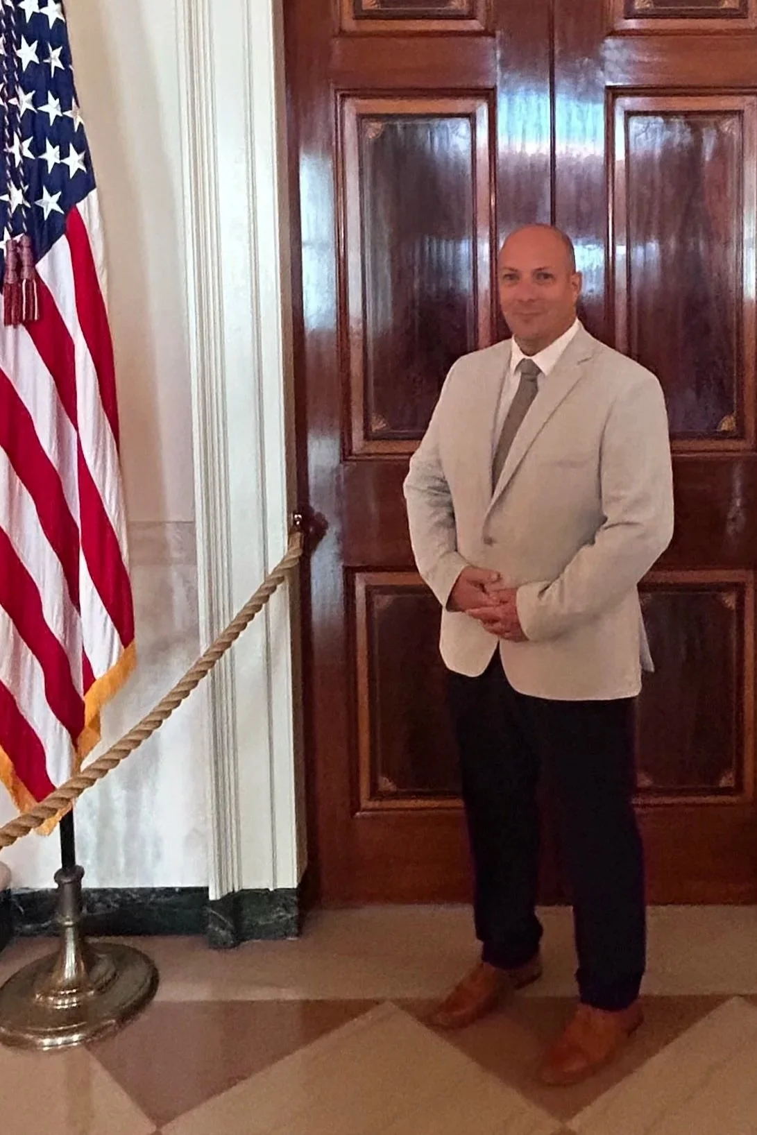 A man in a light-colored suit standing indoors near an American flag and wooden paneling.