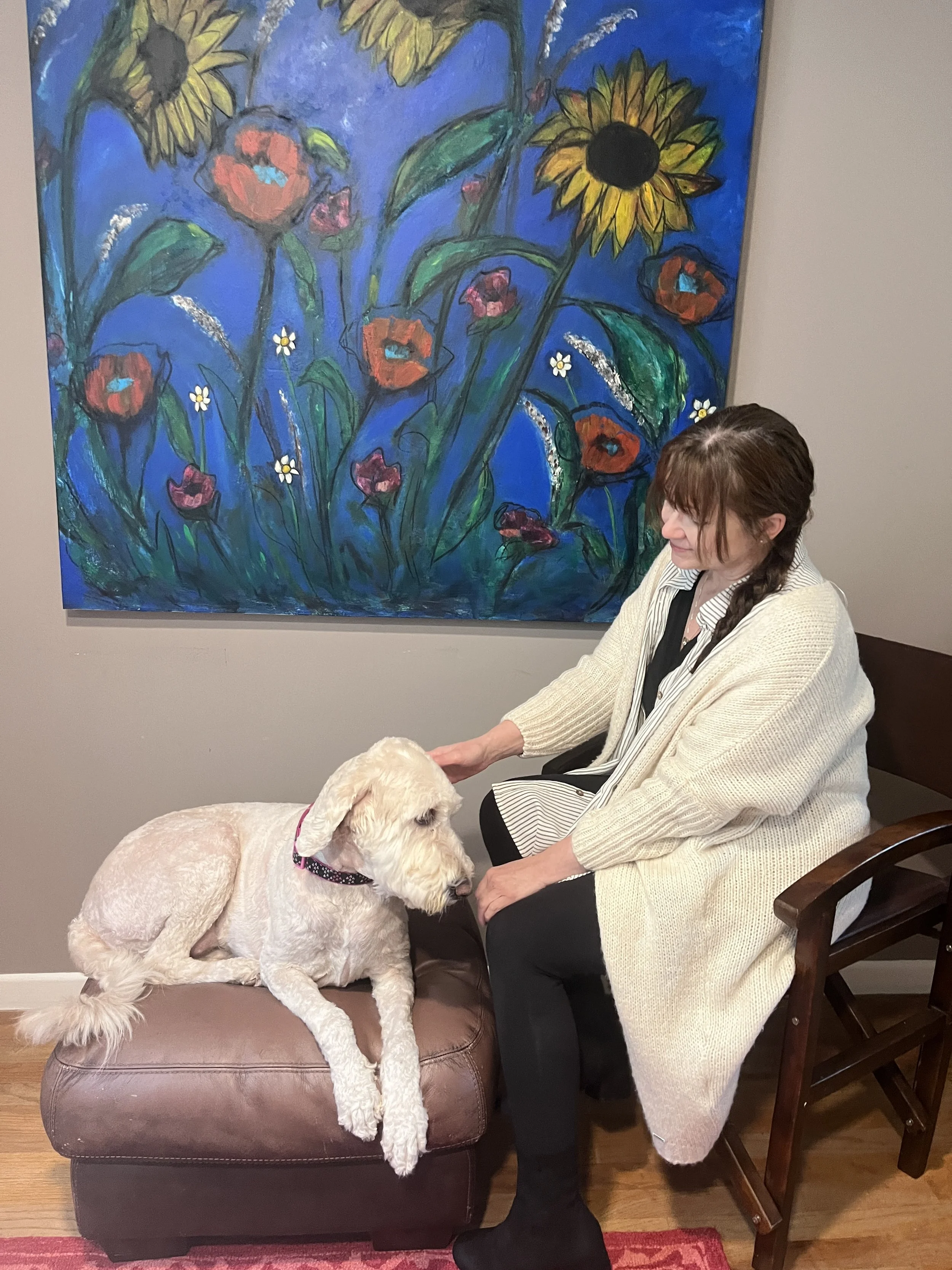 Artist Diane Devine sits with her dog before a painting of sunflowers and poppies she painted.