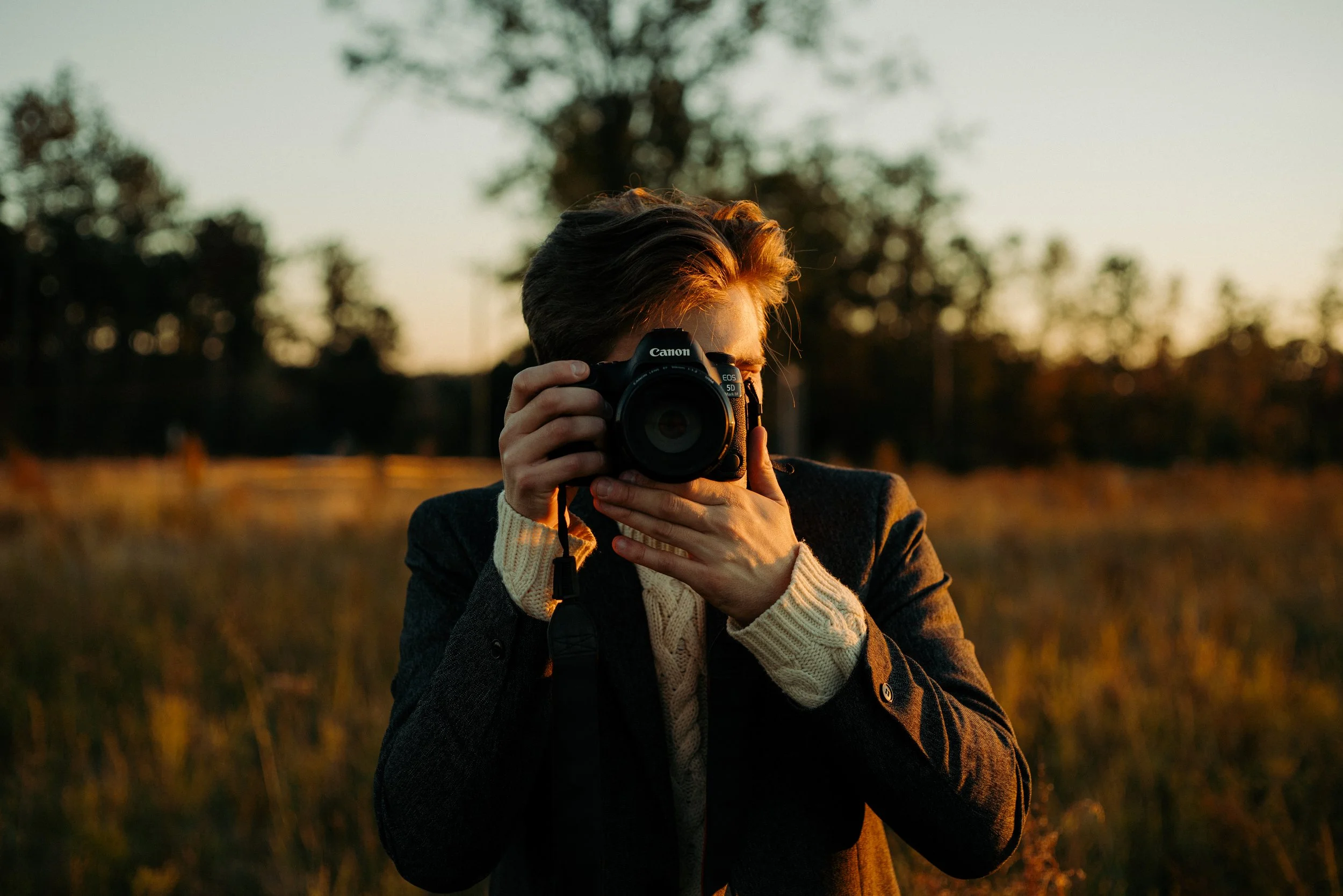A man taking a photo outdoors during sunset, wearing a dark jacket and cream-colored sweater, holding a Canon camera up to their face.