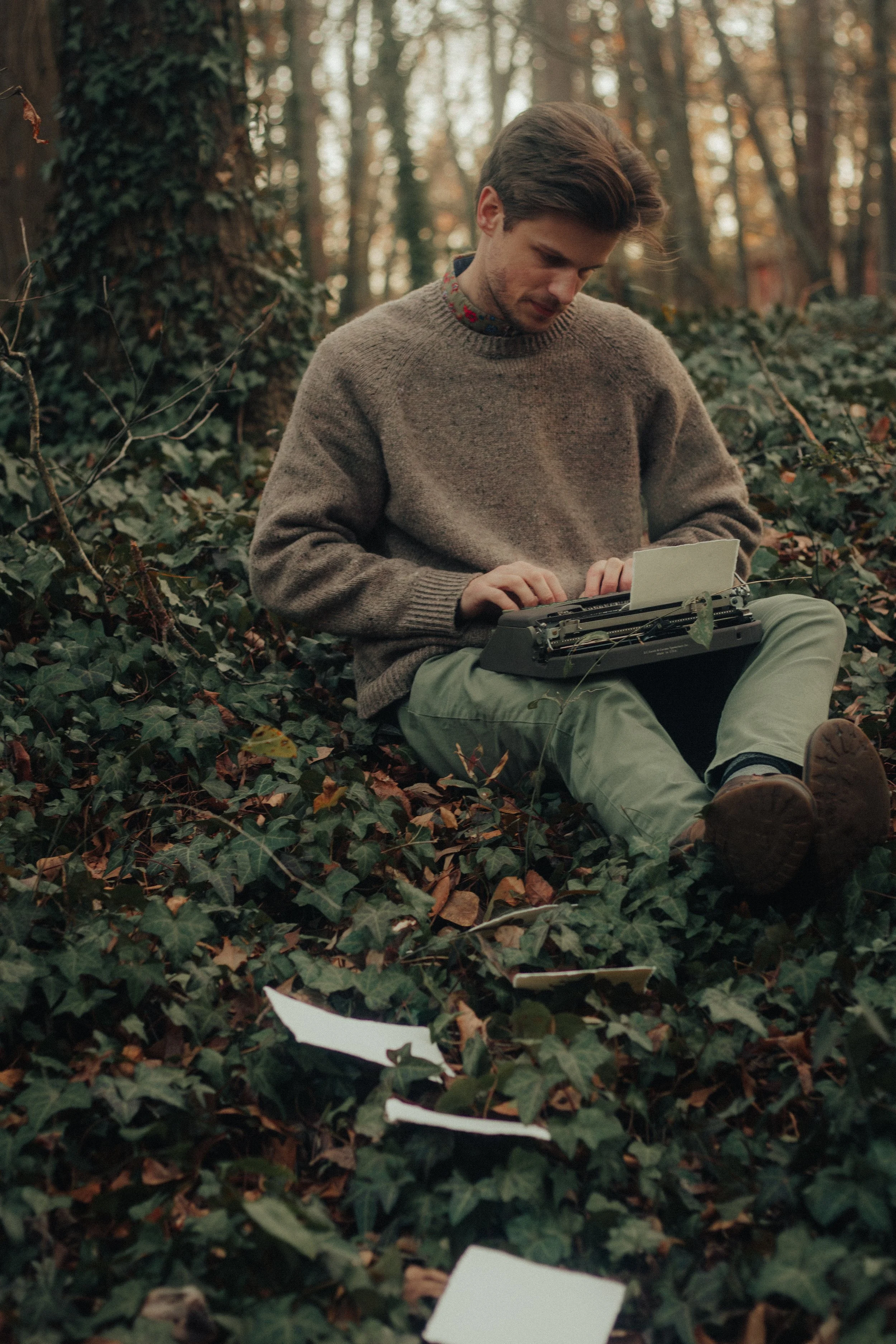 Man (a poet) with a brown sweater, sitting on the ground in a wooded area using an old typewriter, surrounded by fallen leaves and scattered papers.