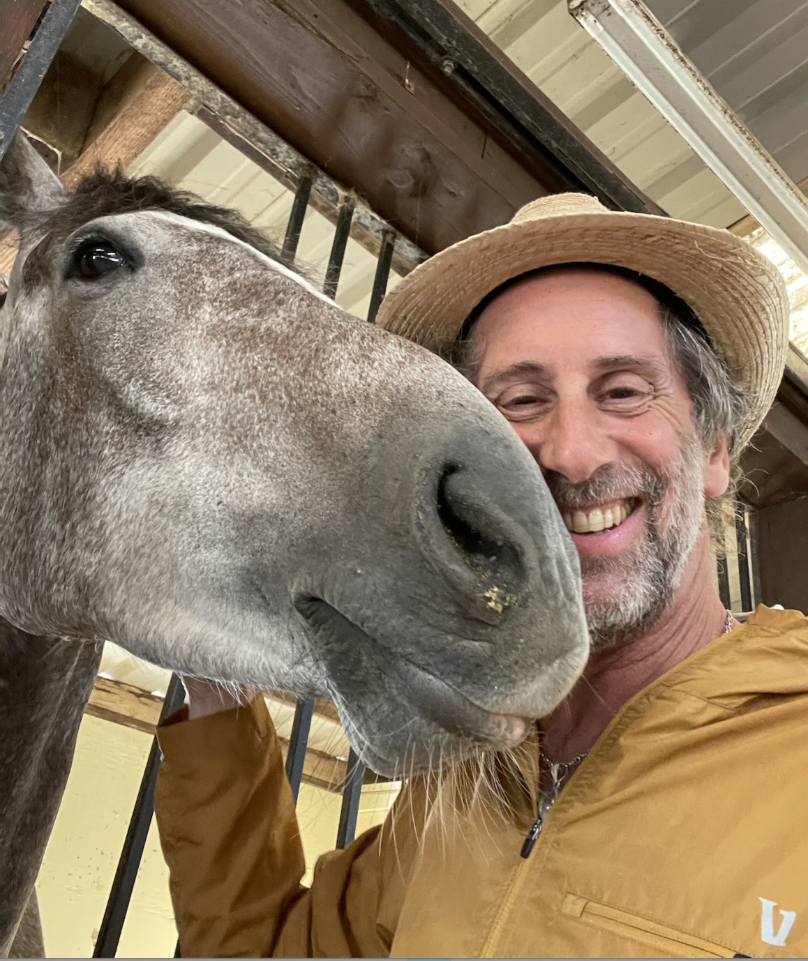 Michael Langer, AMFT, smiling and wearing a straw hat, posing with a gray and white speckled horse, pushing his face into the photo.