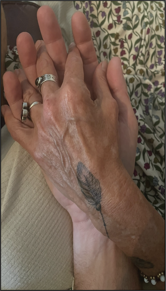 Close-up of two hands holding, conveying a couples love. The one on top is a woman's and has a delicate tattoo of a feather, rings and a bracelet. The other larger hand underneath is a man's, both set against a patterned dress of flowers.