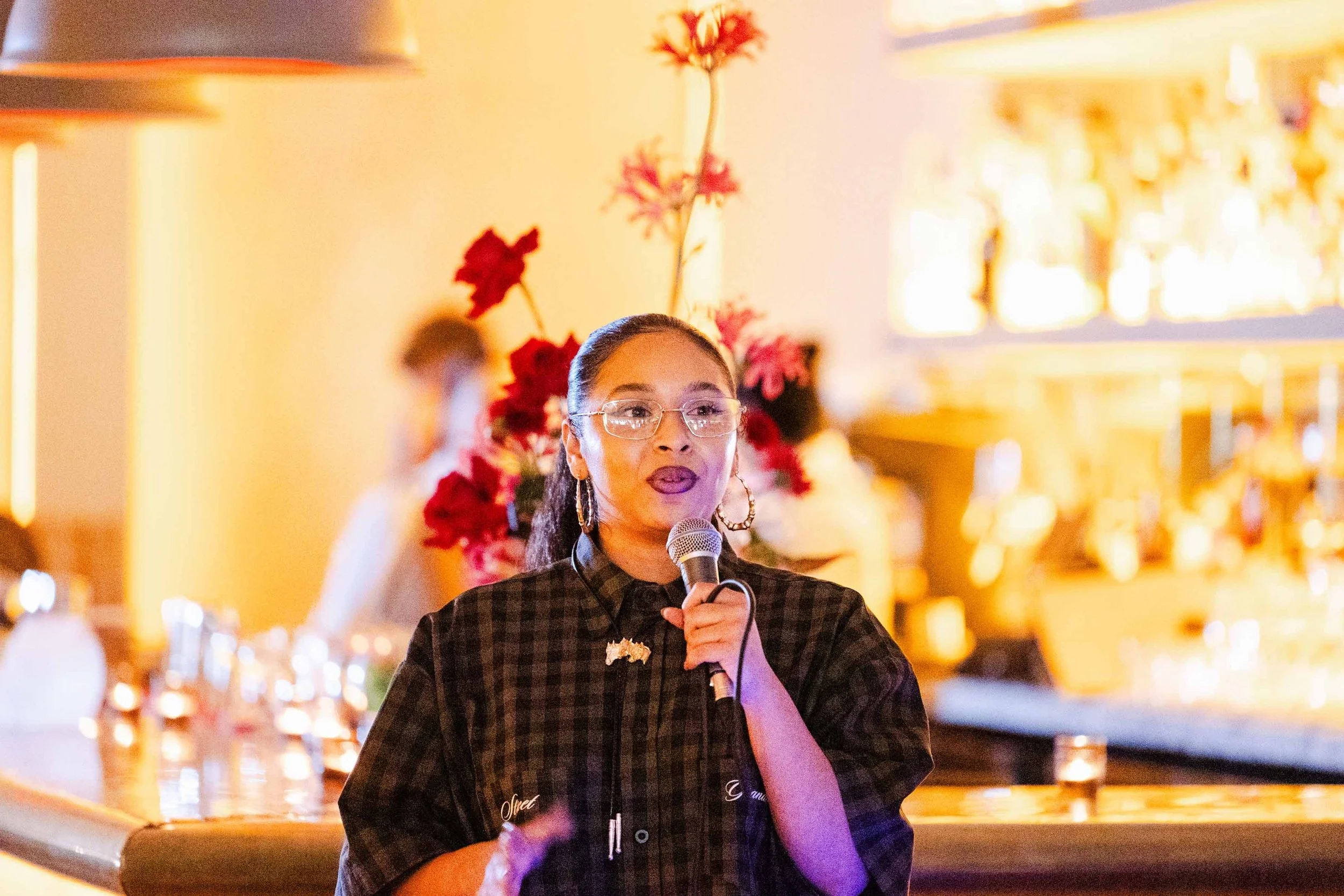 A speaker presents during the Latinas in Beauty launch event in Beverly Hills, with floral arrangements and bar ambiance in the background.